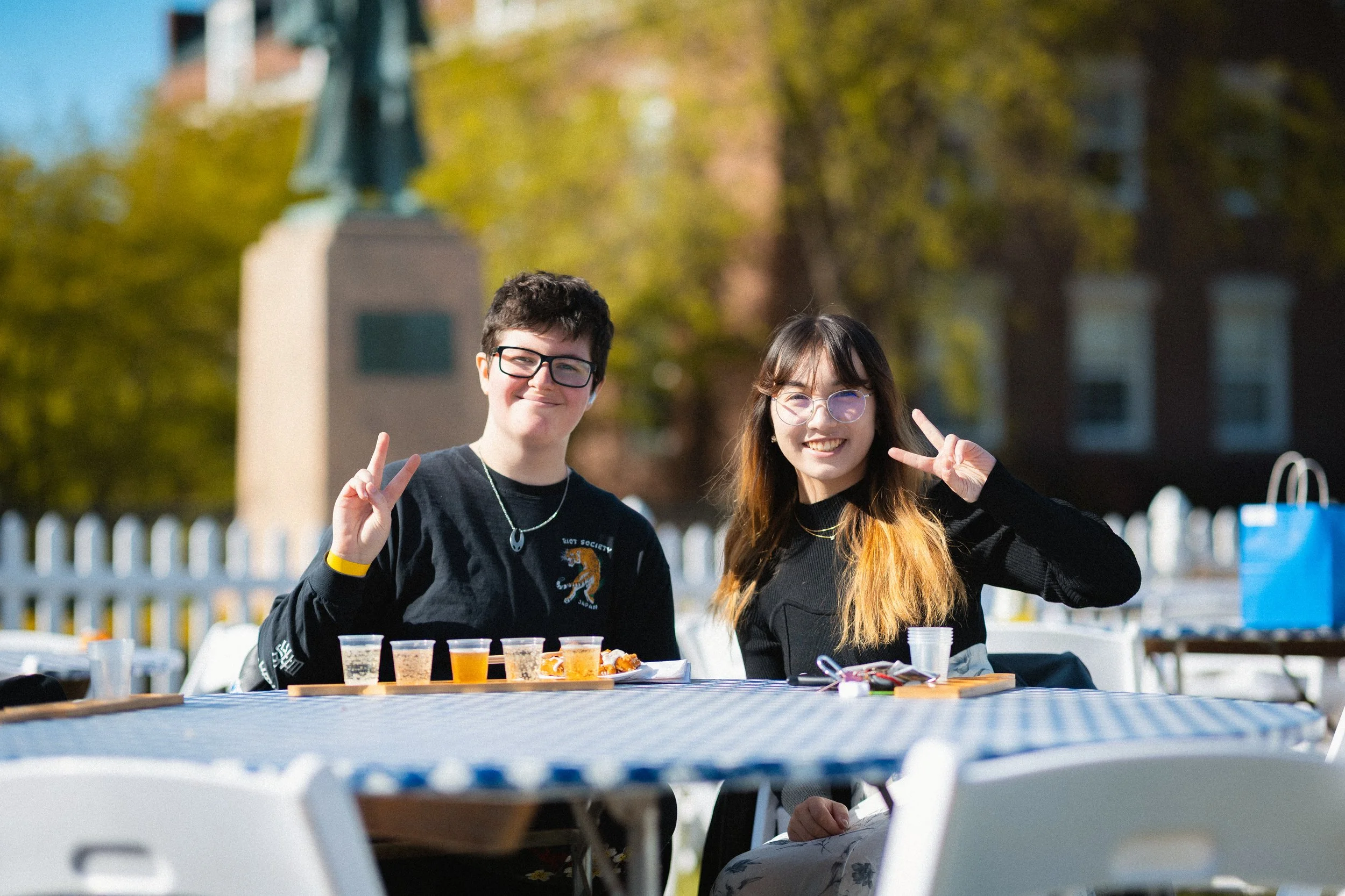 Two smiling teenagers, a boy and a girl, making peace signs with their fingers, sitting at an outdoor table with small cups of beverages and snacks, during a sunny day in a park with trees and a white picket fence in the background.