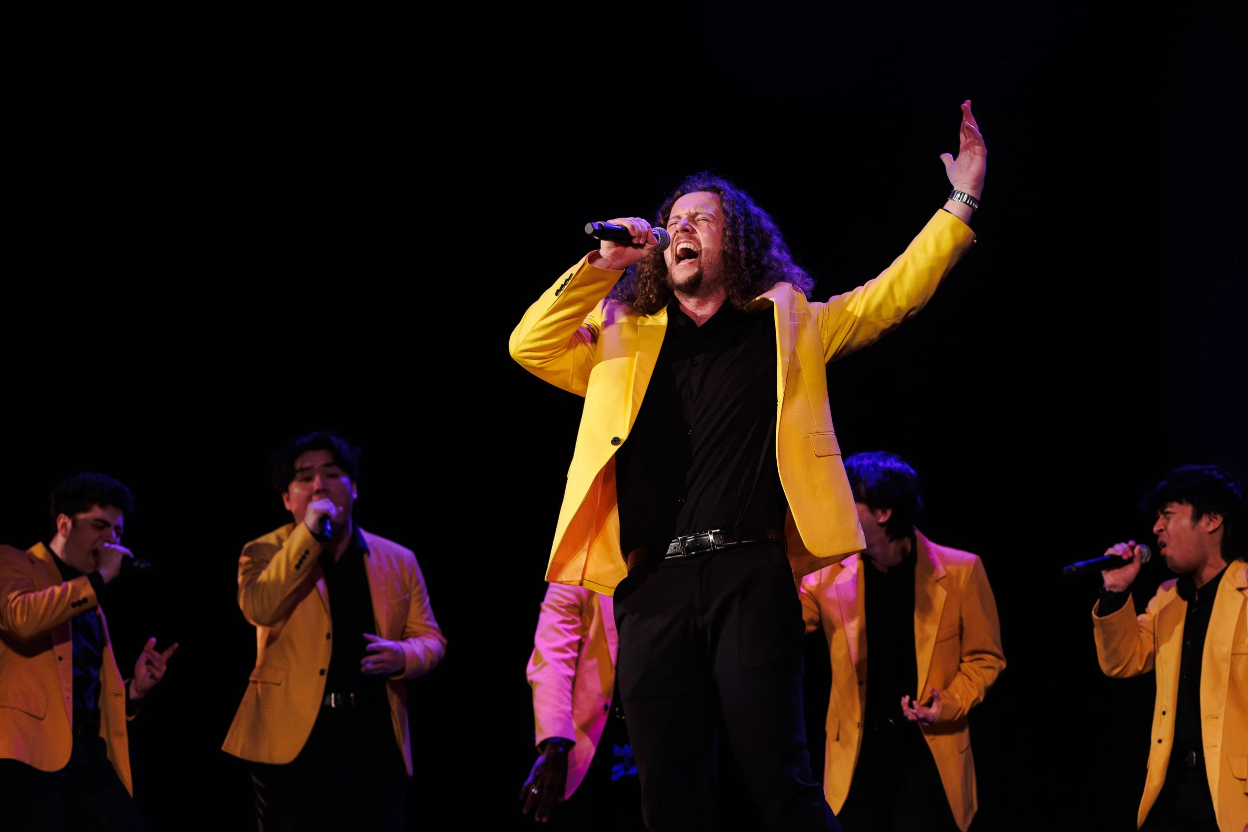 A male singer in a yellow blazer and black shirt passionately singing into a microphone, with his right hand raised, surrounded by band members also dressed in yellow blazers, performing on stage against a black background.
