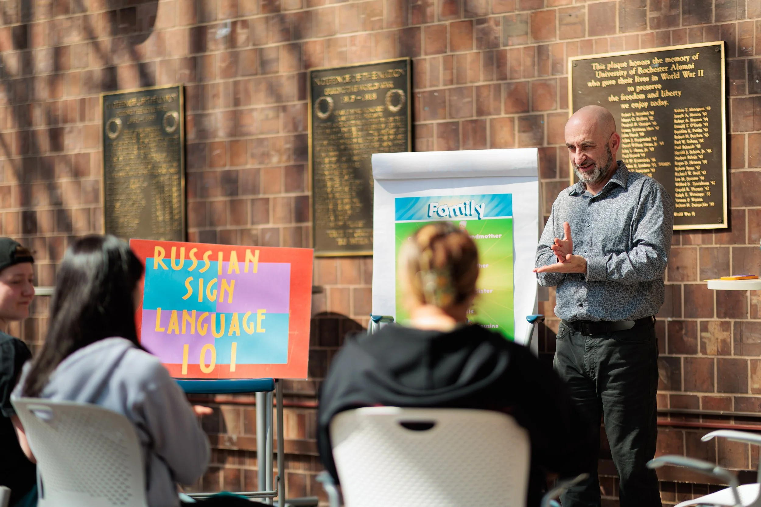 A man giving a presentation outdoors in front of a brick wall, with posters on the wall and audience members seated in chairs.