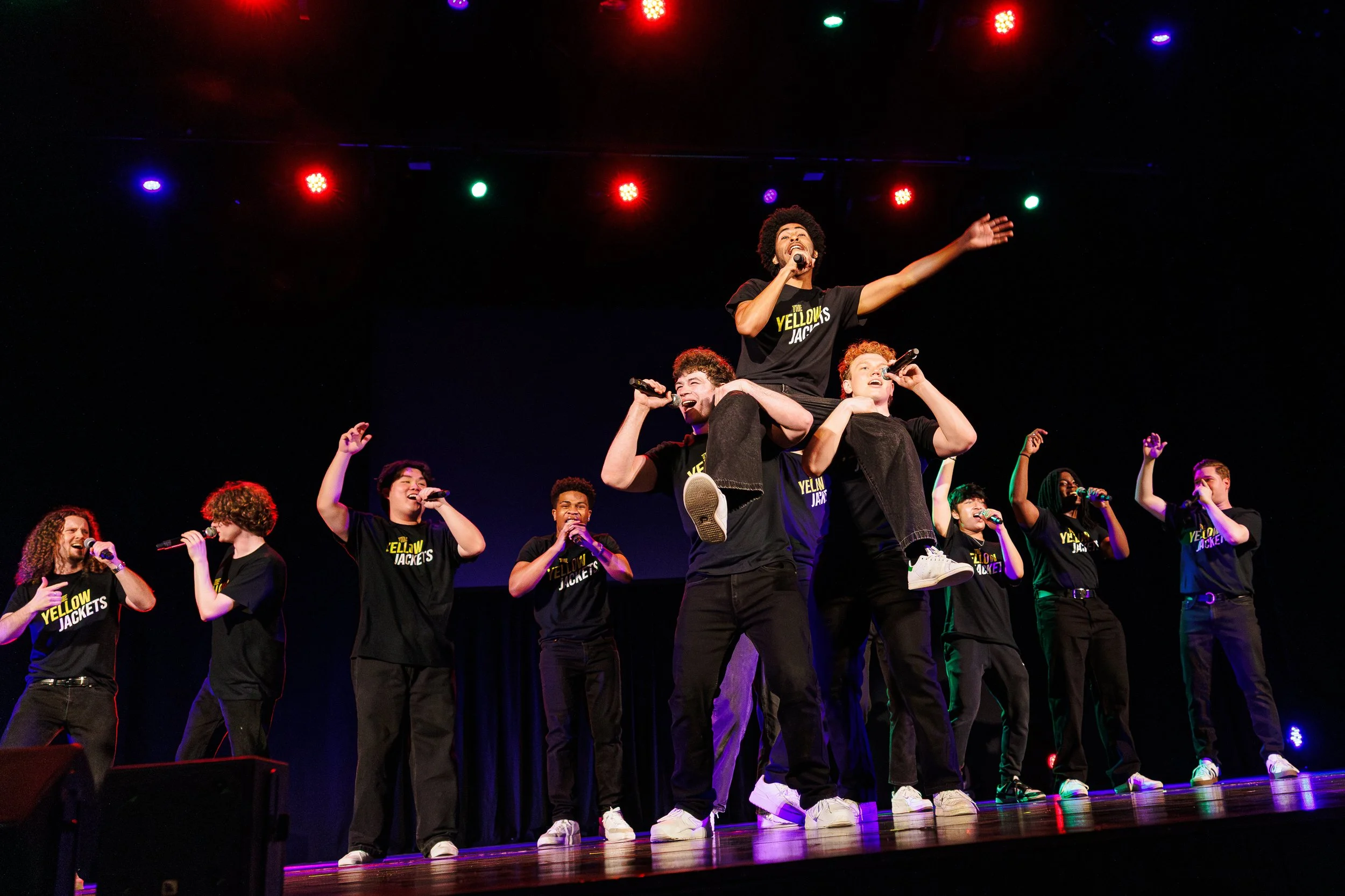 A group of young people on stage performing at a concert or event. One individual is sitting on another person's shoulders, holding a microphone and wearing a T-shirt that reads 'The Yellows Jackets.' The others are also singing into microphones, rai