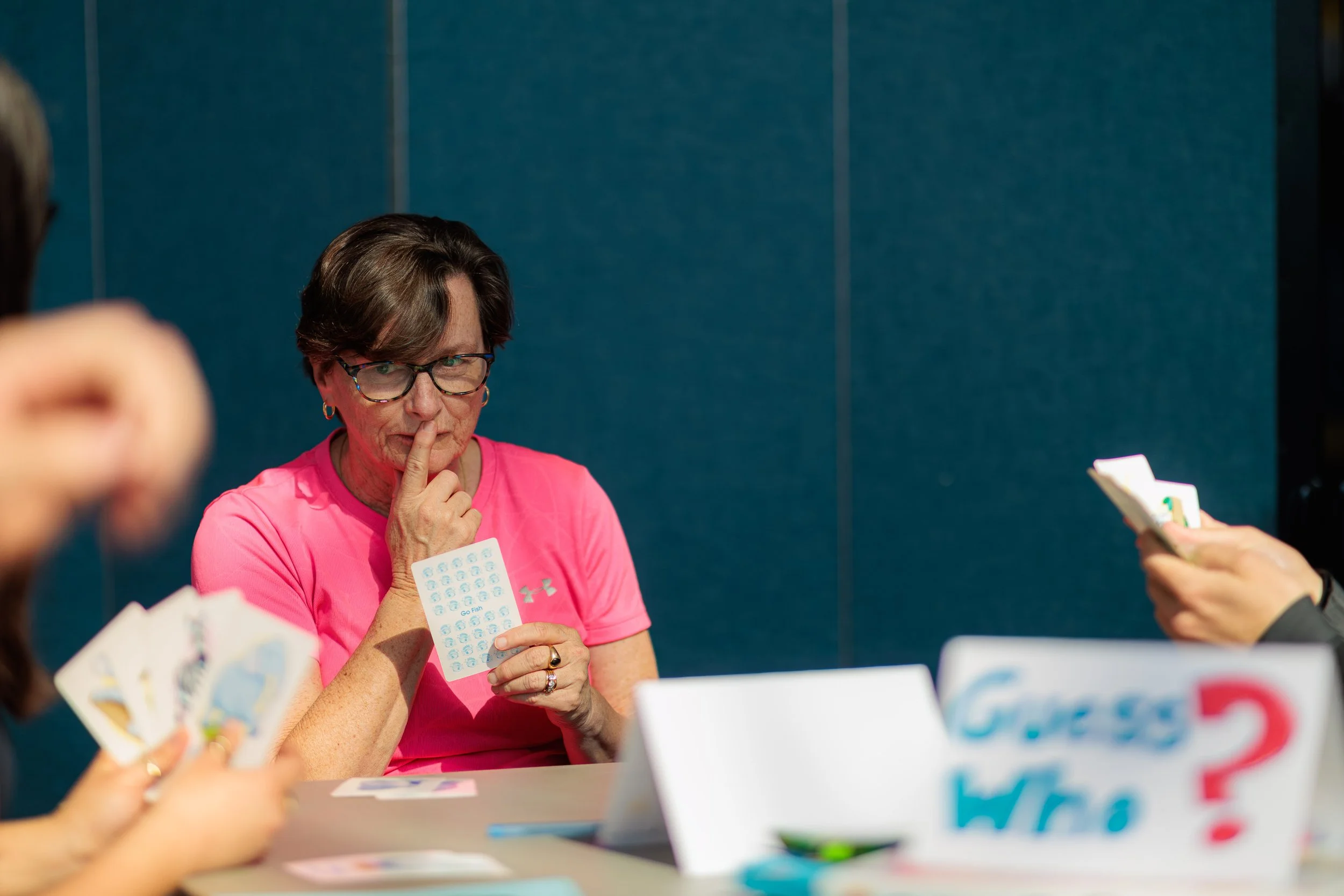 A woman with short brown hair, glasses, and earrings wearing a pink shirt is holding a bingo card and looks thoughtful, touching her chin. She is surrounded by other people holding bingo cards, in a room with a blue wall.