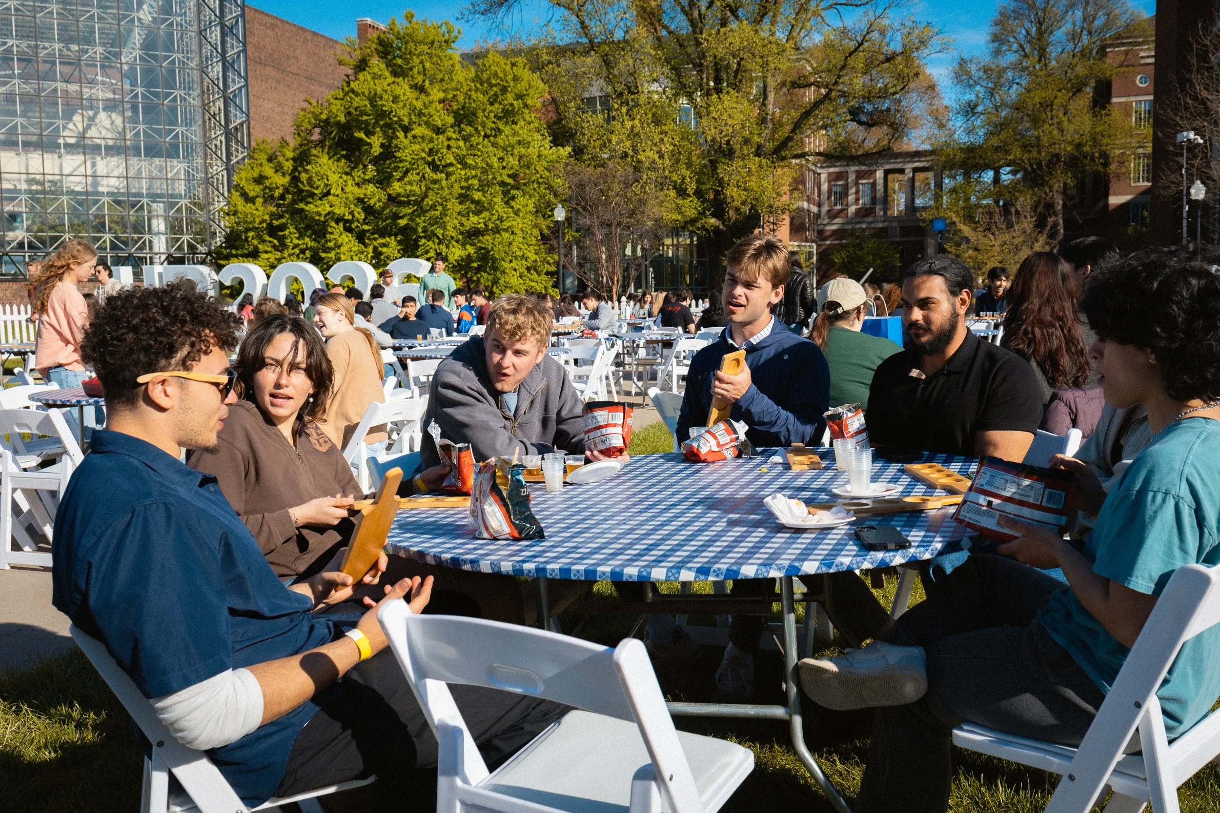 Students sitting at round tables outdoors during a sunny day, engaging in conversations and eating snacks, with trees and a building in the background.