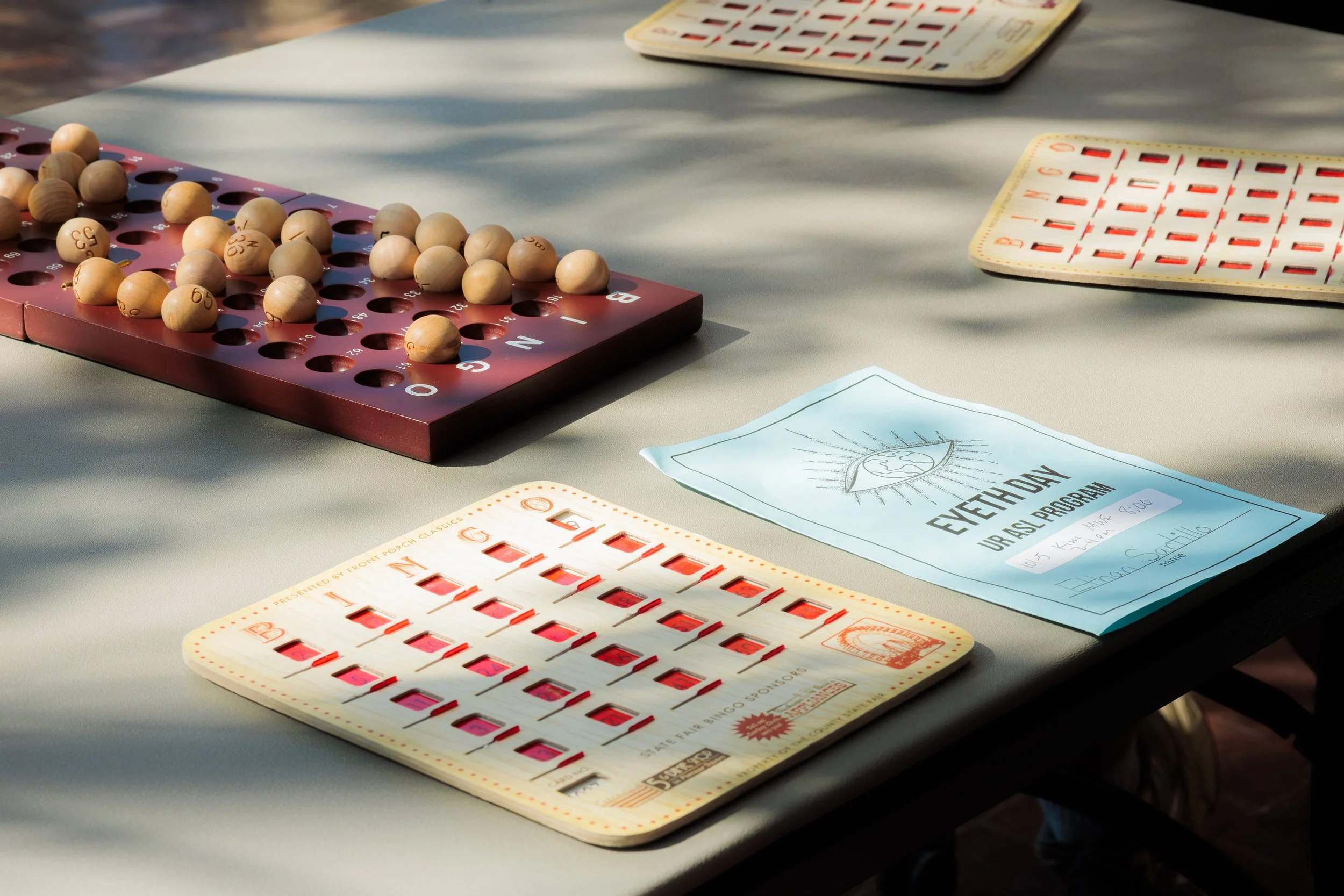 A Bingo game setup on a table featuring wooden numbered balls in a pink and brown container, Bingo cards, and a handout with an event day program.