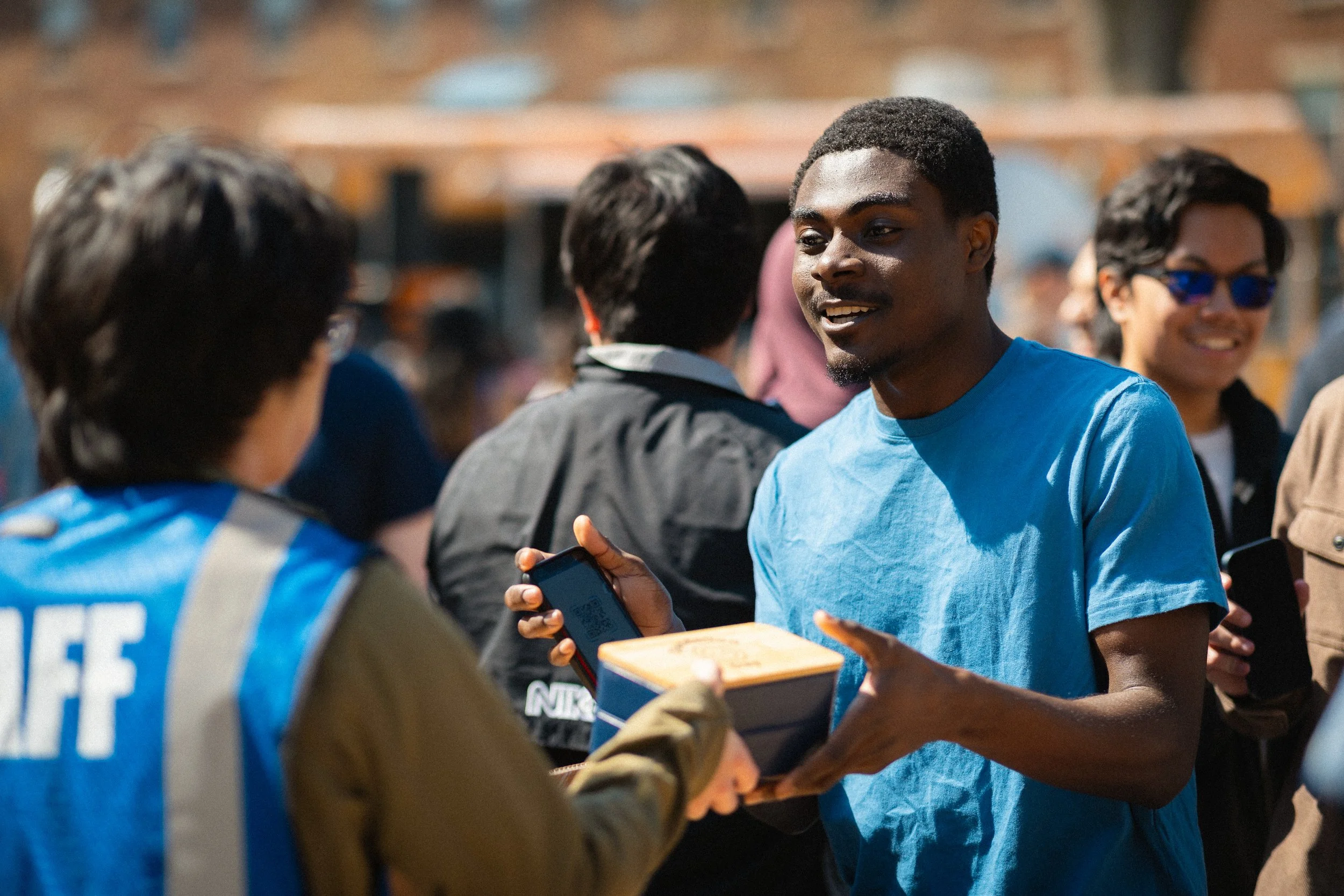 A young man wearing a blue shirt and holding a box and a phone, smiling and talking to other people at an outdoor event during daytime, with several people in the background.