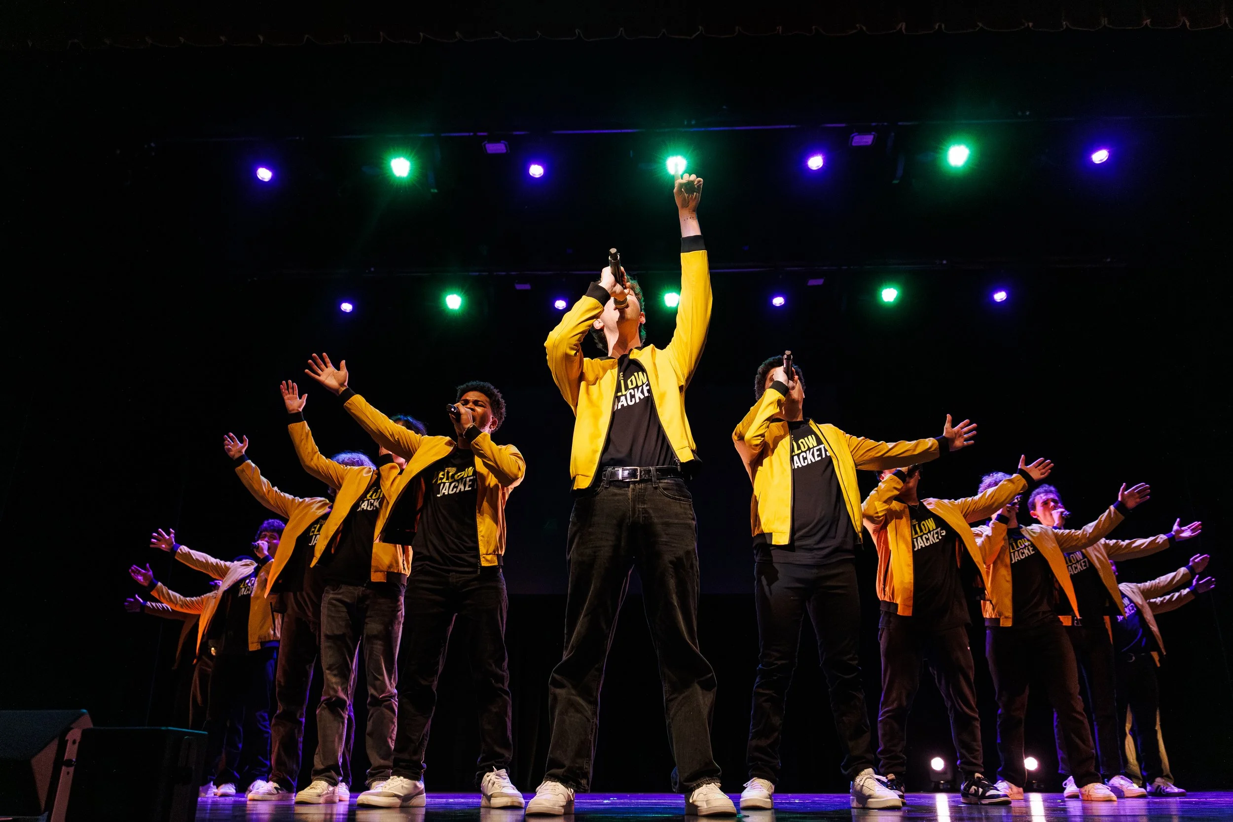 A group of performers dressed in yellow jackets with black shirts underneath are dancing on stage. The stage is lit with colorful lights, and the performers have their arms raised in the air. The performers seem to be part of a choreographed dance ro