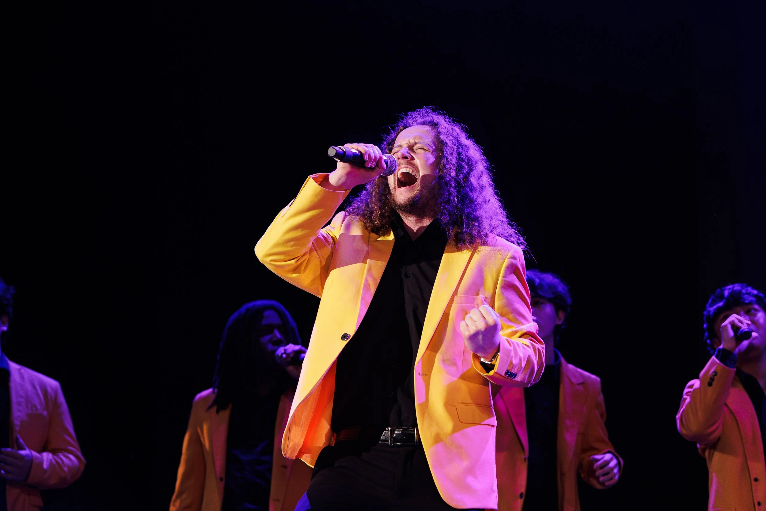 A man with curly hair singing passionately into a microphone on a stage, wearing a bright yellow blazer, with backup singers in yellow blazers in the background.