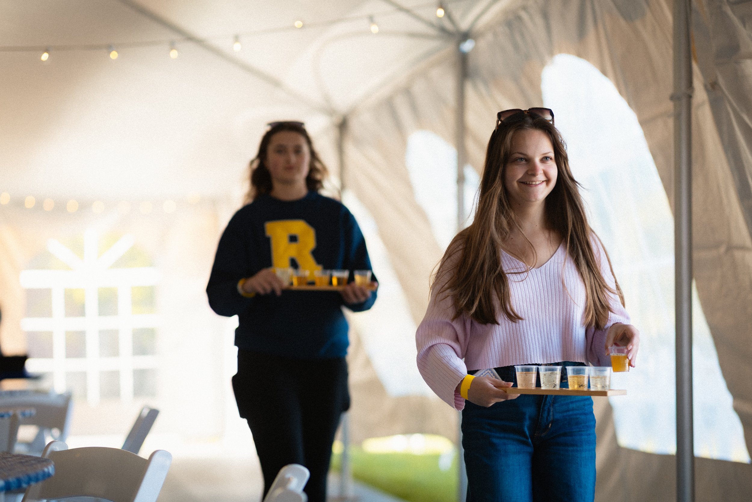 A young woman smiling and serving drinks on a tray at an outdoor event under a canopy, with a young woman in a dark sweater carrying drinks in the background.