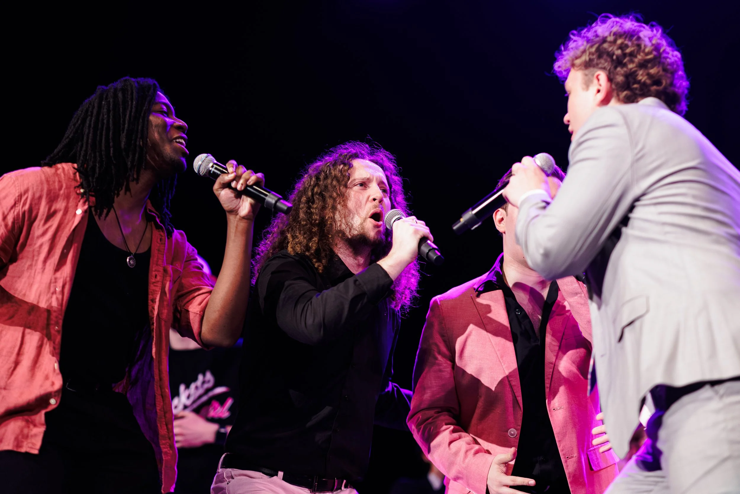 Group of people singing into microphones on stage during a performance, with a dark background and purple stage lighting.