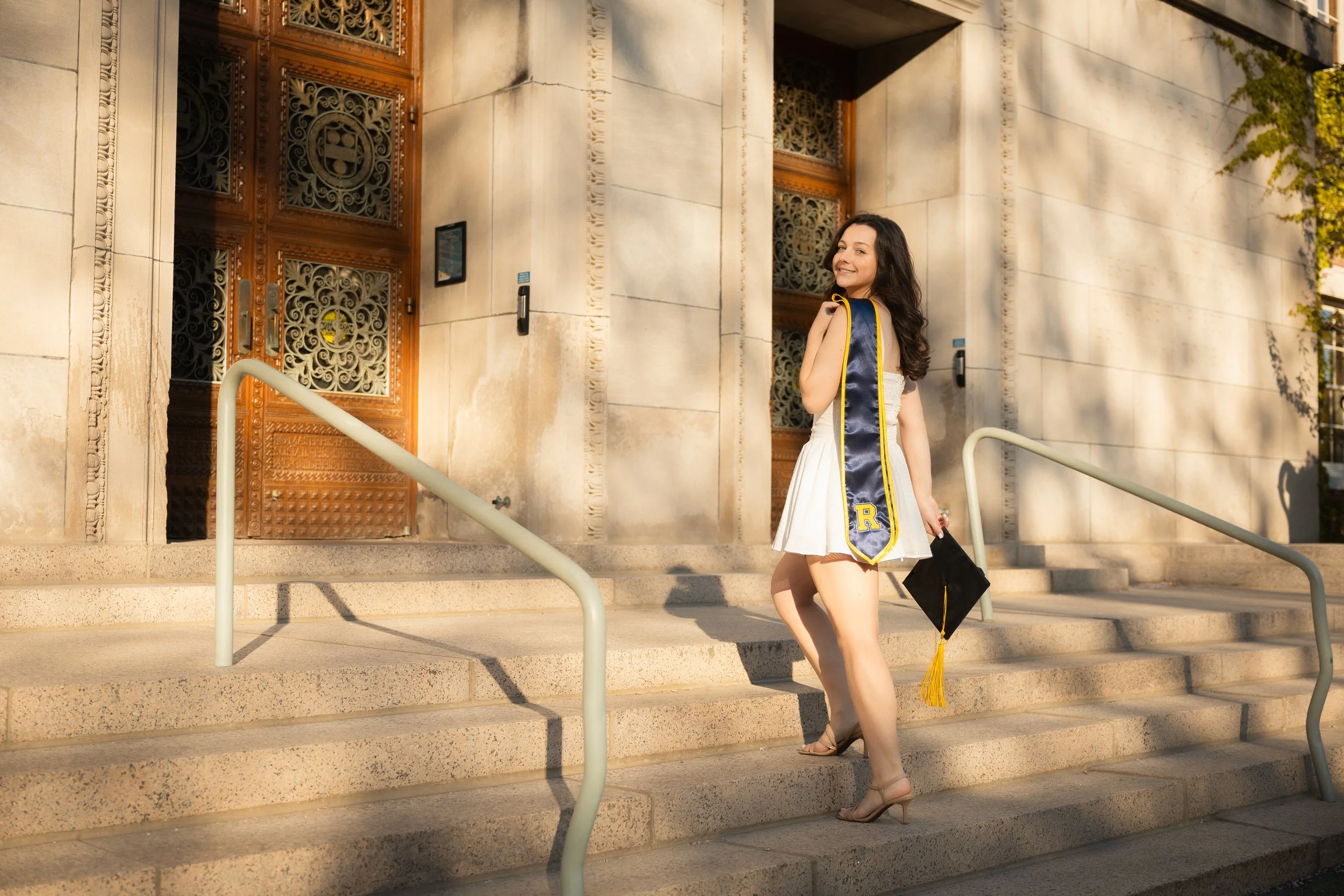 Young woman in a white dress and high heels standing on stone steps outside a building with ornate wooden doors, holding a diploma with a gold tassel and smiling at the camera.