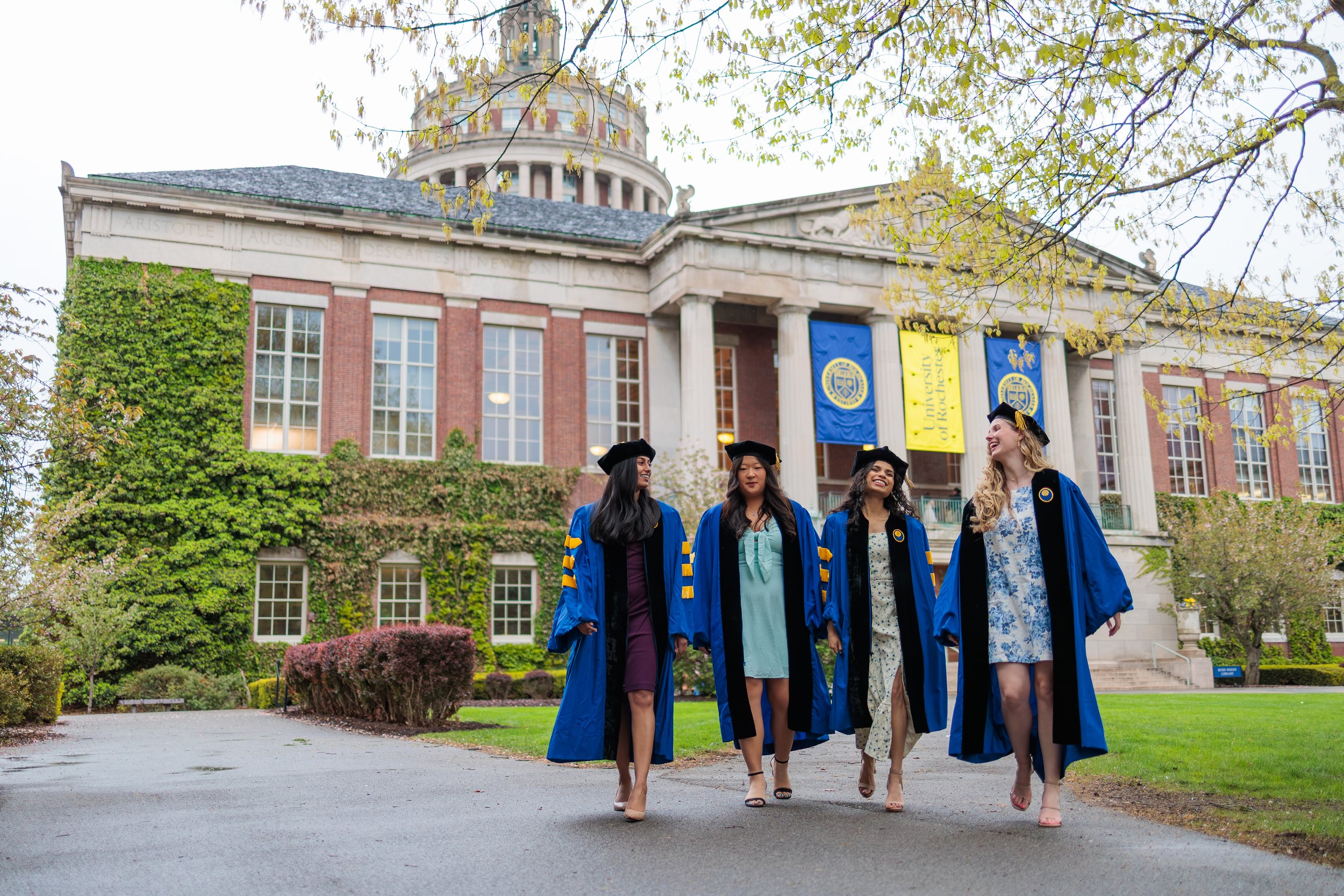 Four women in graduation caps and gowns walking on a path in front of a university building with columns and flags. The women are smiling and dressed in formal attire.