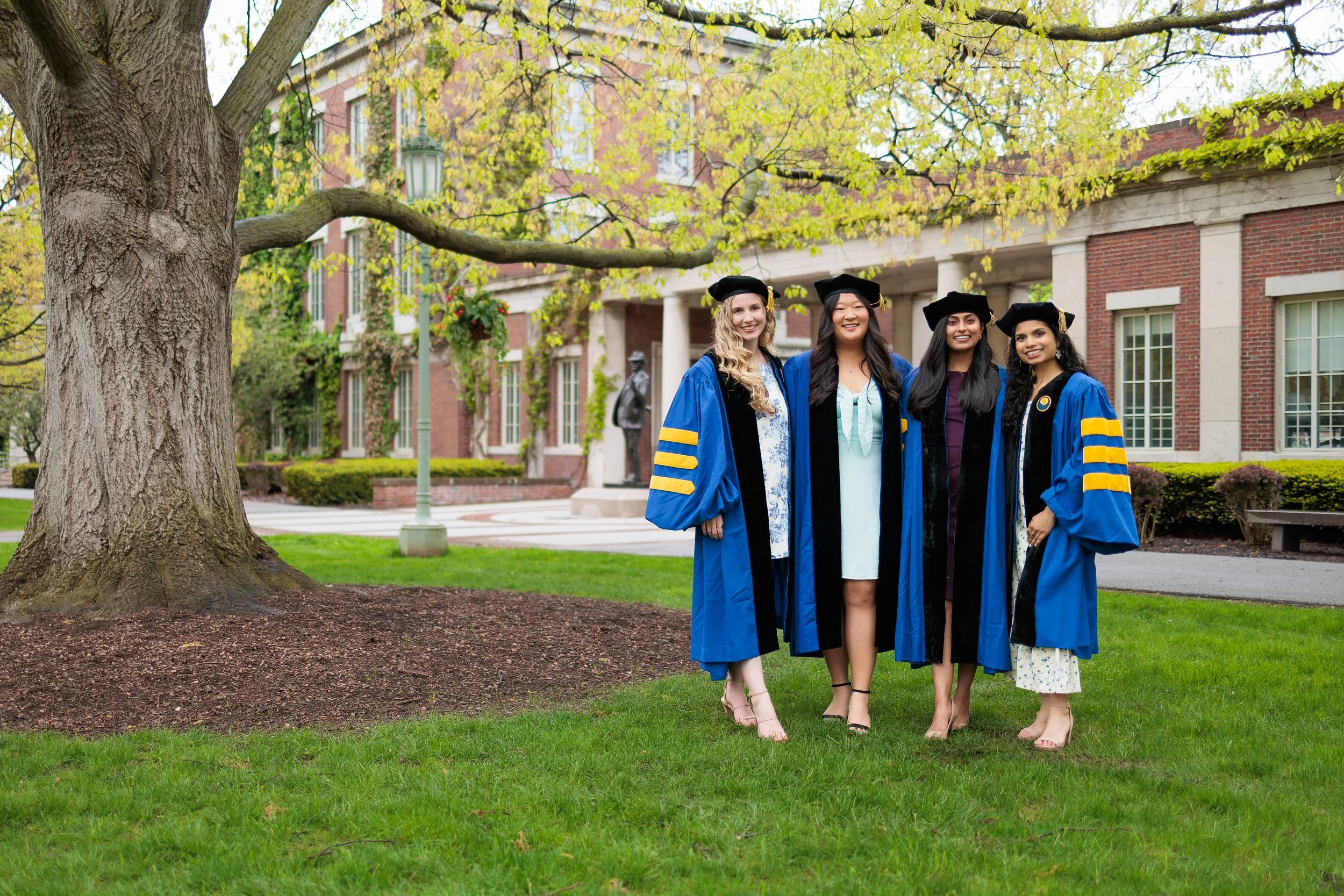 Four women dressed in blue graduation gowns and caps standing outdoors in front of a brick building. They are smiling and posing for the photo beneath a large tree with green leaves.