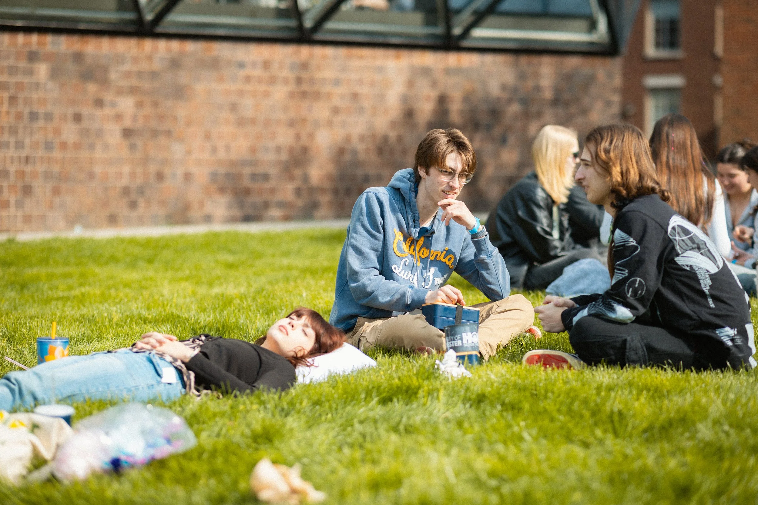 Group of teenagers sitting on the grass outside near a brick building, some chatting and one girl lying down with her eyes closed. There are snacks and drinks around them, and the scene appears to be a casual outdoor gathering.