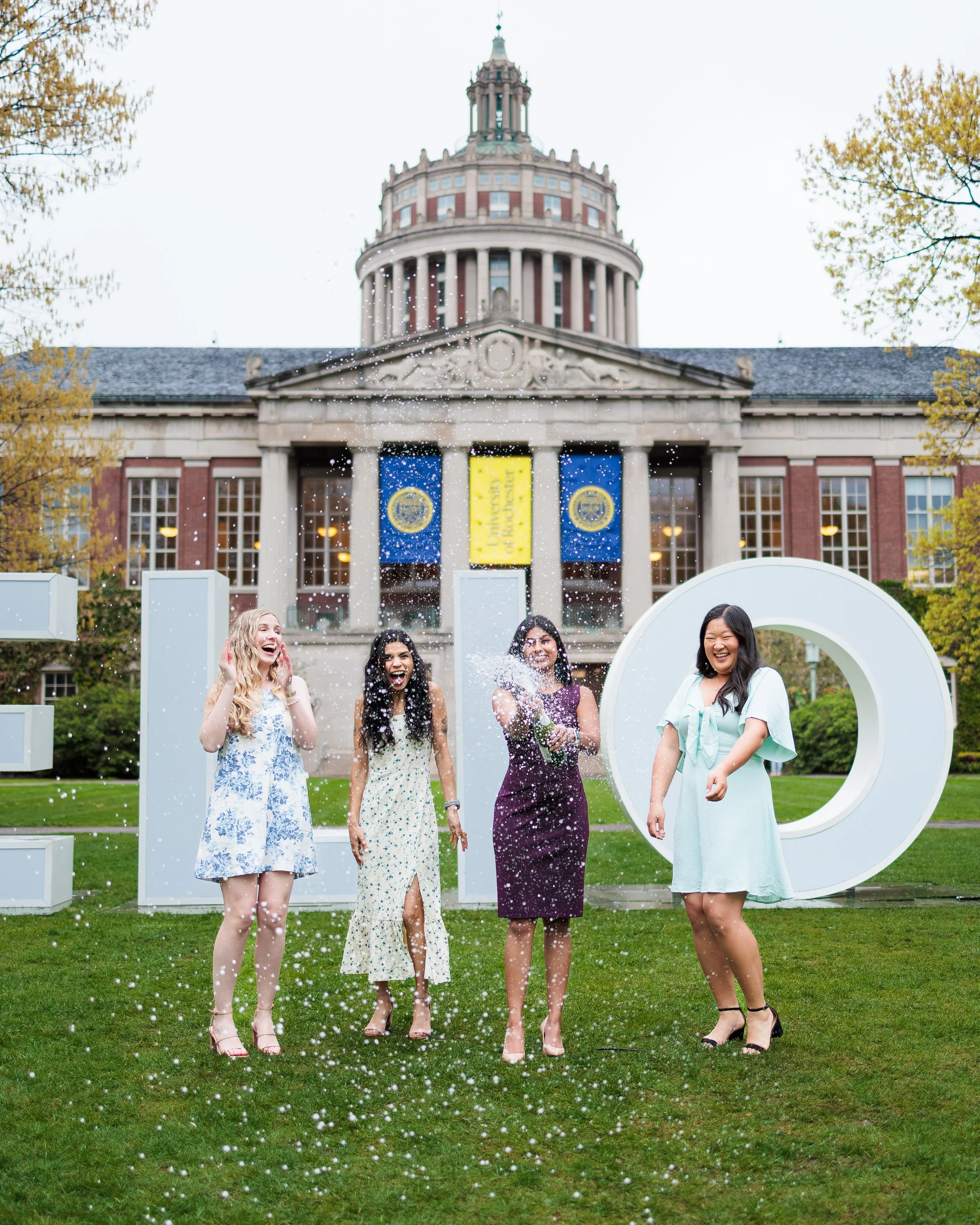 Four women standing outdoors on a grass lawn, celebrating with confetti, in front of large white decorative letters, with a historic building and trees in the background.