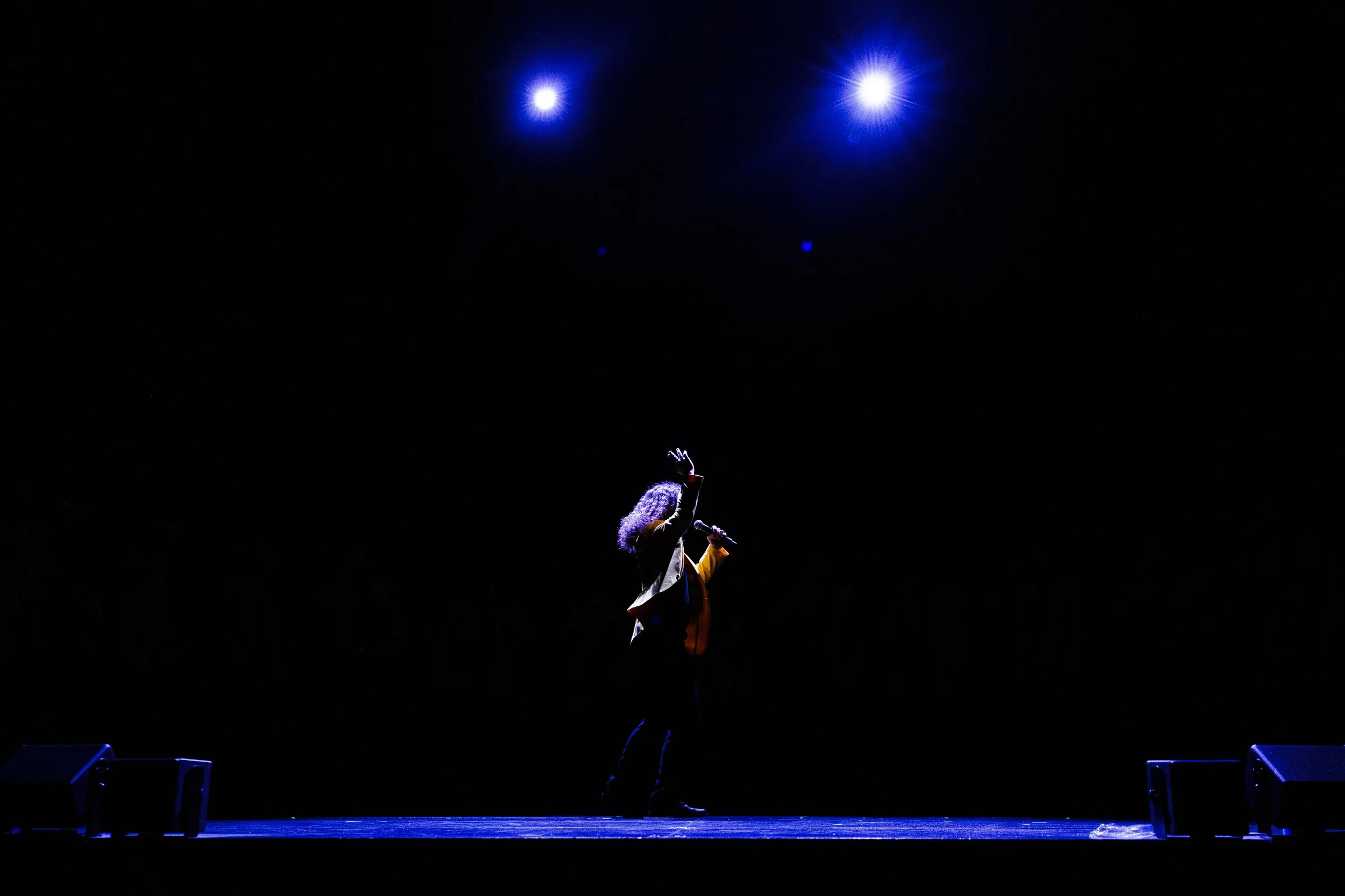 A person standing on a stage holding a microphone, with the spotlight shining on the person, in a dark setting.