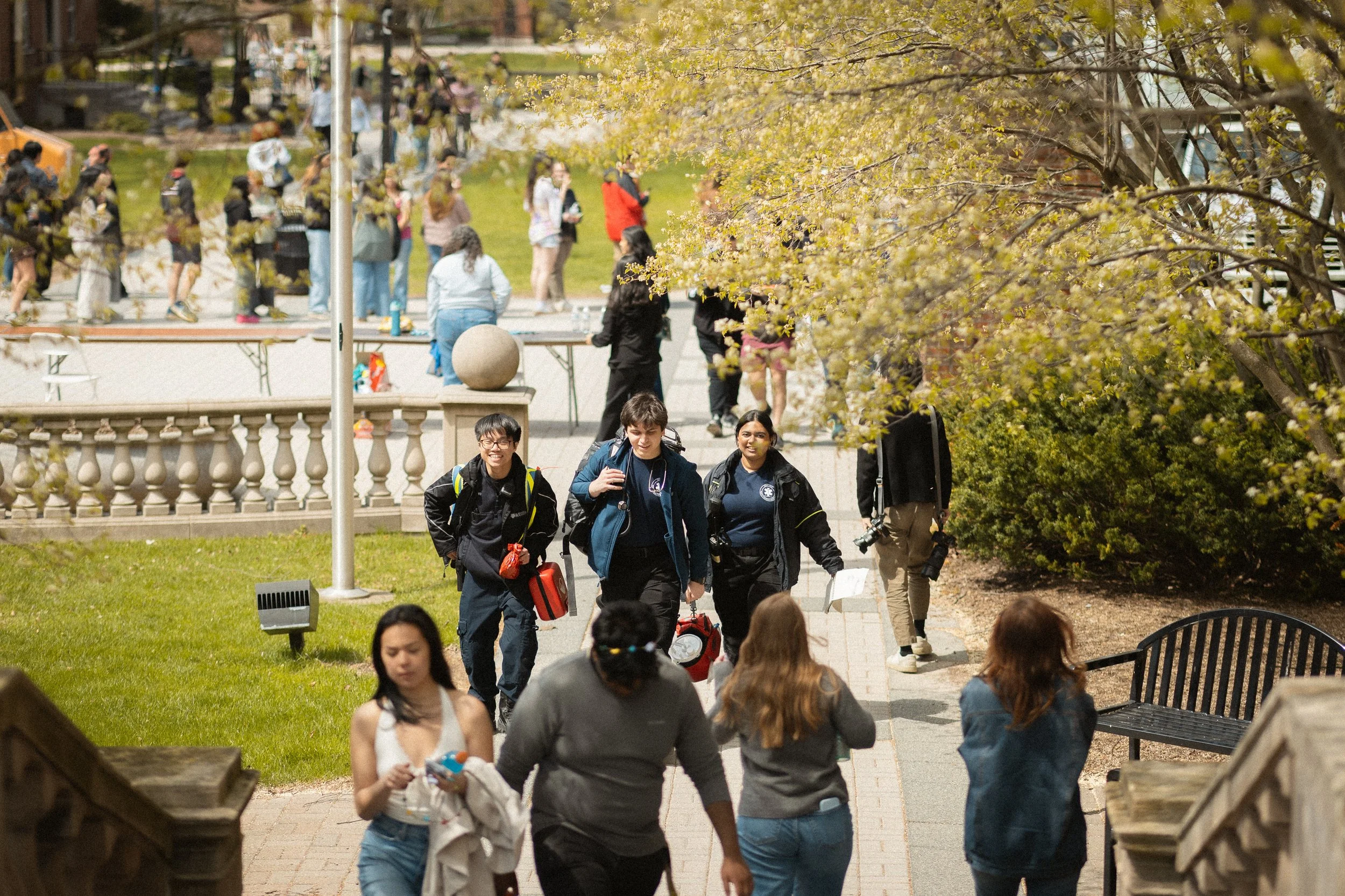 People walking and gathering in an outdoor area with trees and benches, some standing in line or chatting, in a park or campus setting.