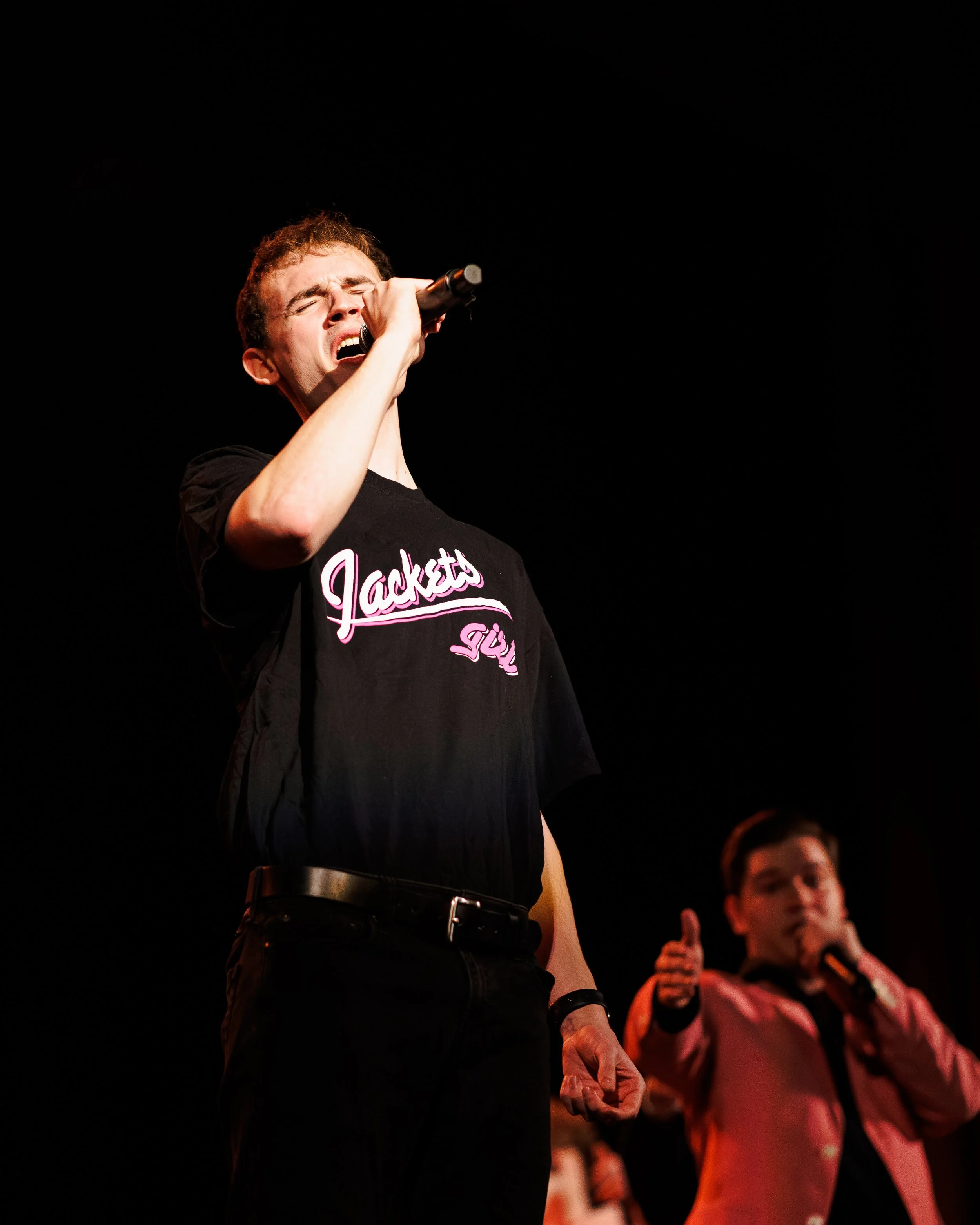 A man passionately singing into a microphone on stage with a dark background, wearing a black t-shirt that says "Jackets". In the background, a woman in a pink blazer appears to be gesturing with her hand.
