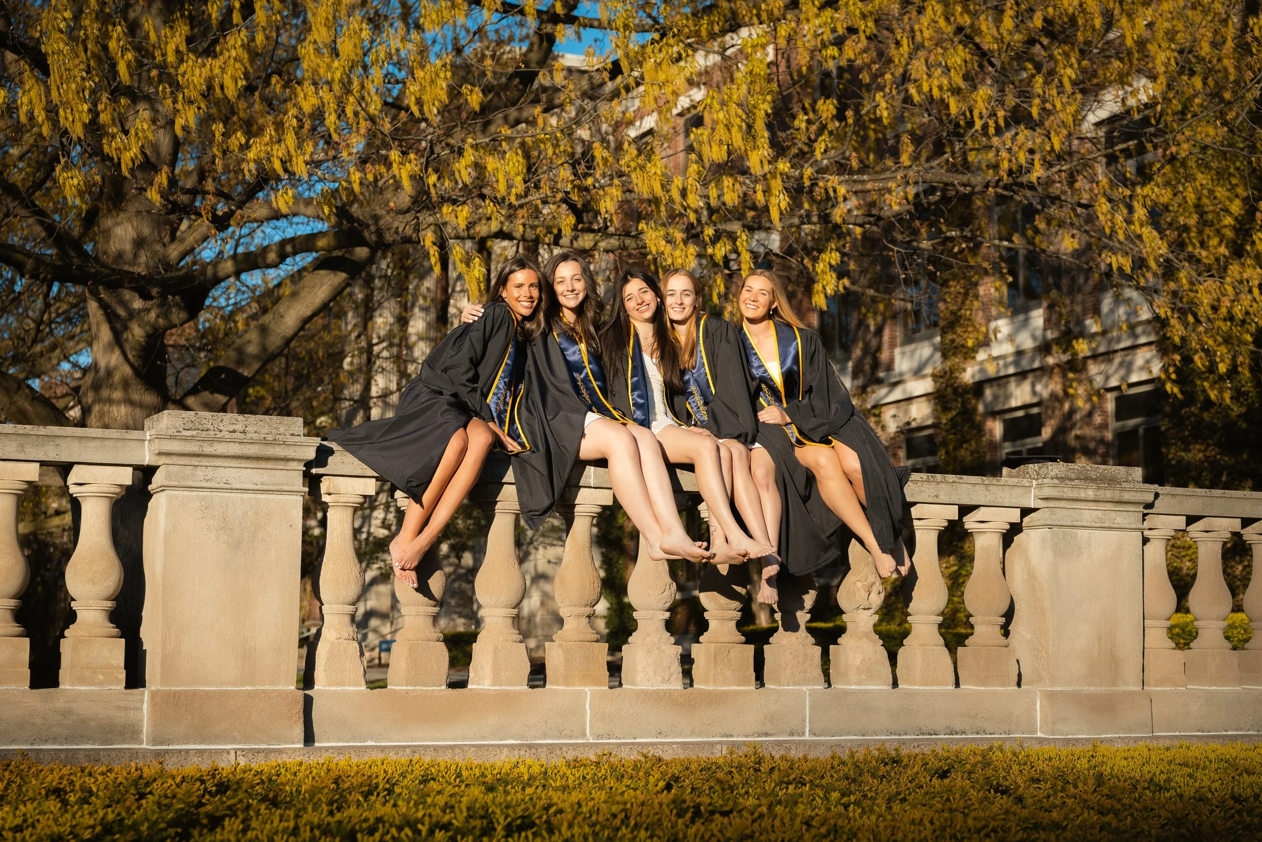 Group of five young women in graduation gowns sitting on a stone balustrade outdoors during autumn, with yellow leaves on trees in the background.