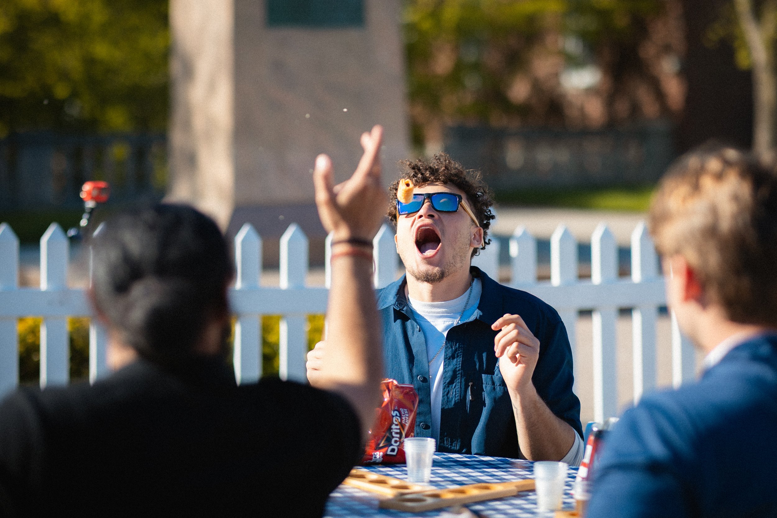 Man with sunglasses and curly hair shouting or talking animatedly to a group of people at an outdoor event, with a table and white picket fence in the background.