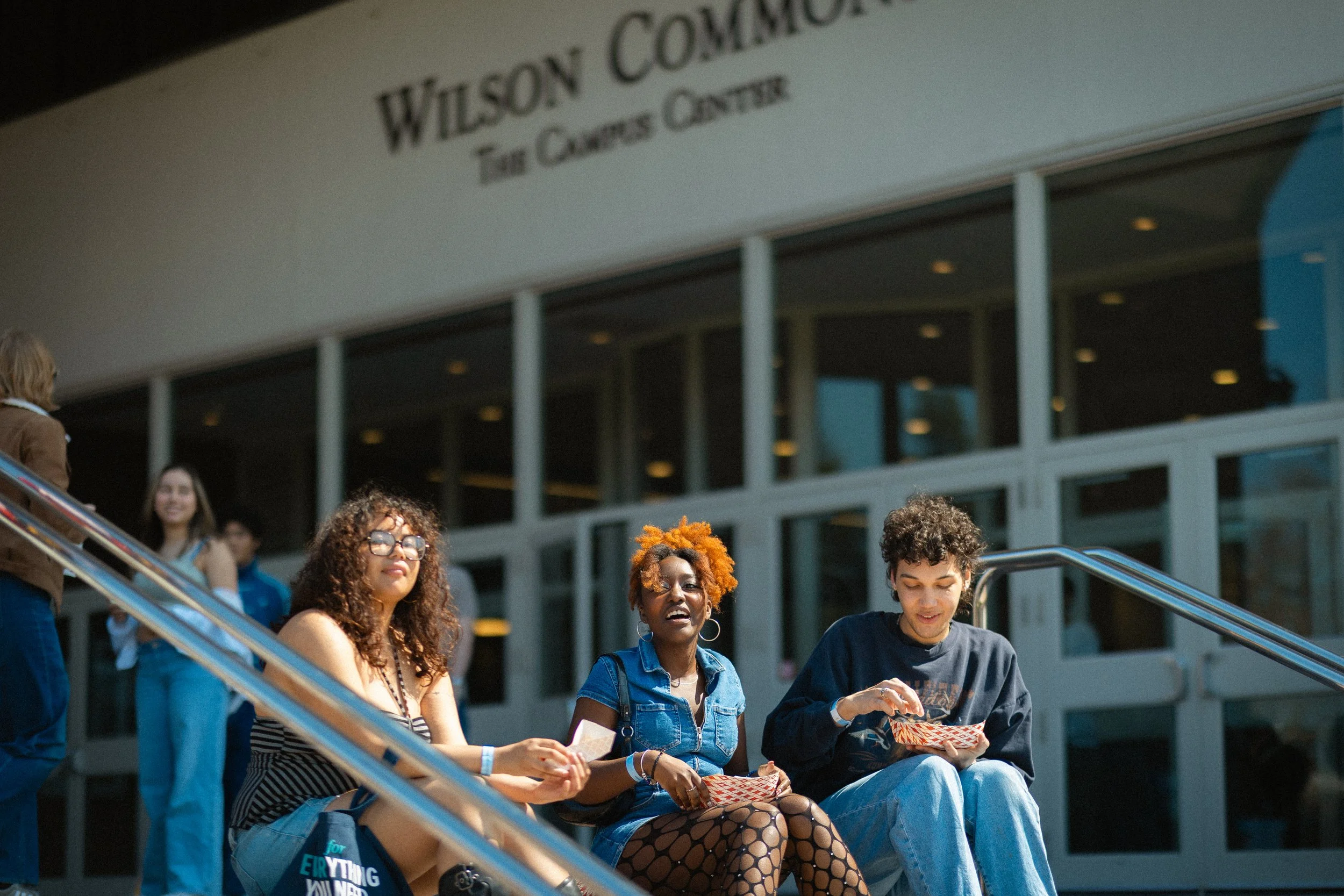 Group of four young people sitting on outdoor steps in front of a building named Wilson Center, with others standing and walking nearby. They appear engaged, some looking at phones or talking.