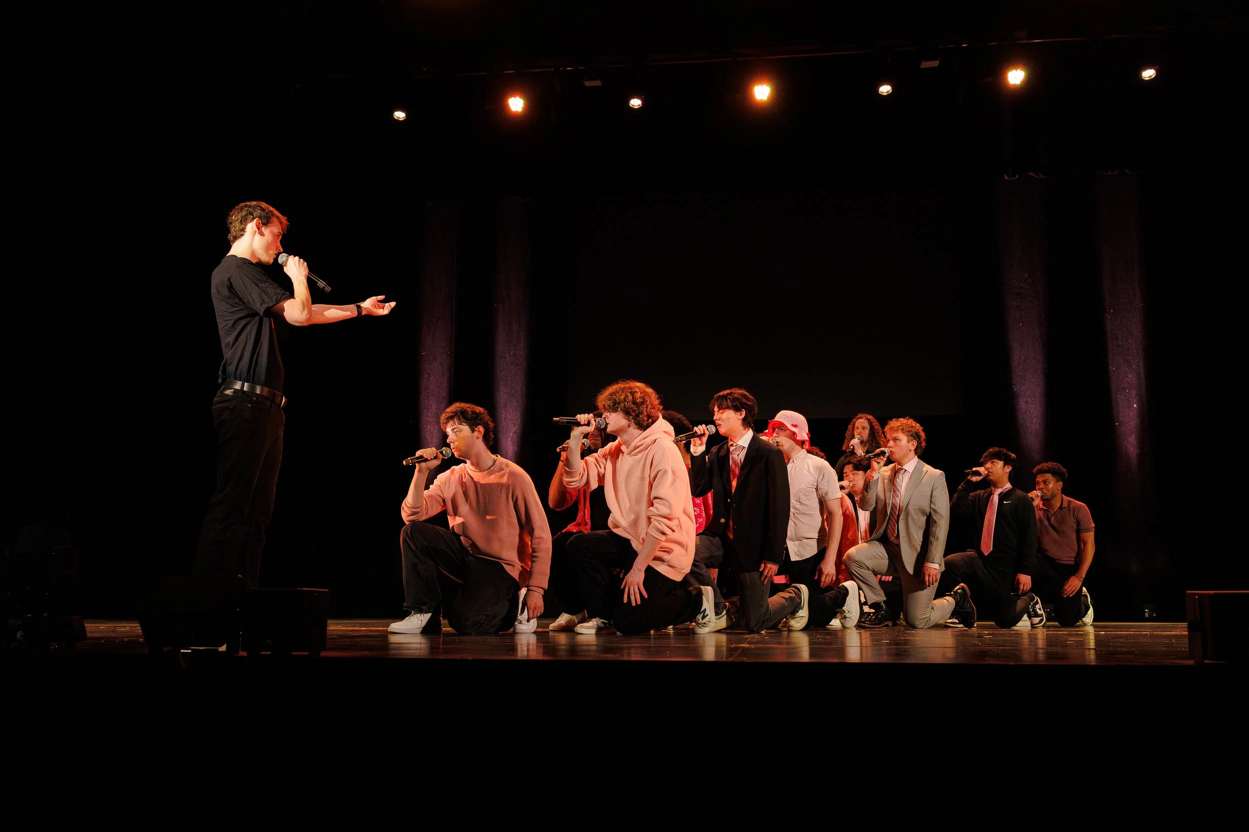 A stage performance with a young man speaking into a microphone while a group of young people kneel and sit in front of him, holding microphones, in a theater with stage lighting.