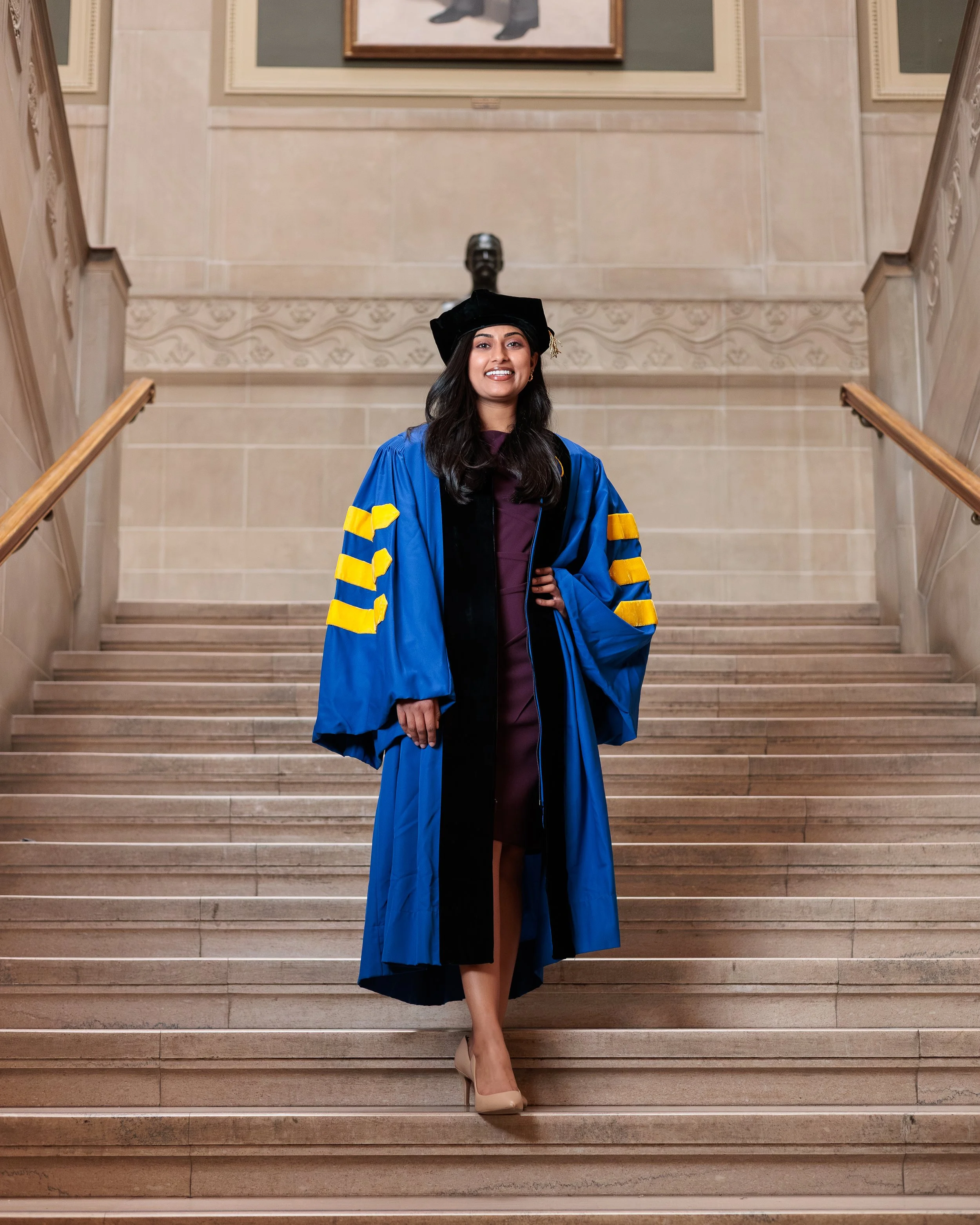 A young woman in a graduation regalia standing on a staircase, smiling, with a painting hanging on the wall behind her.