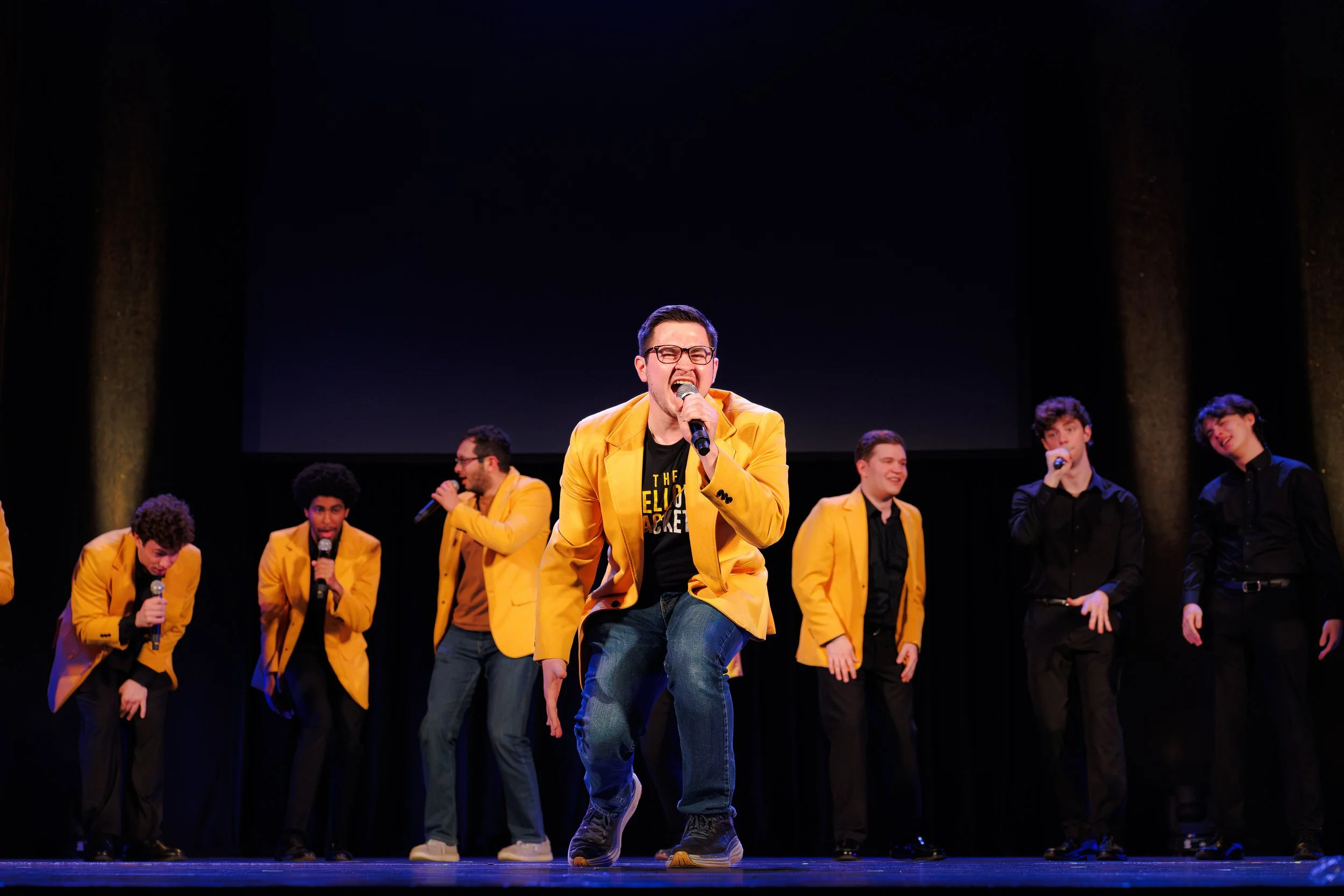 A group of young men performing on stage, with one man singing into a microphone at the center, wearing a yellow blazer and glasses, surrounded by others dressed in black and yellow, with a dark background.