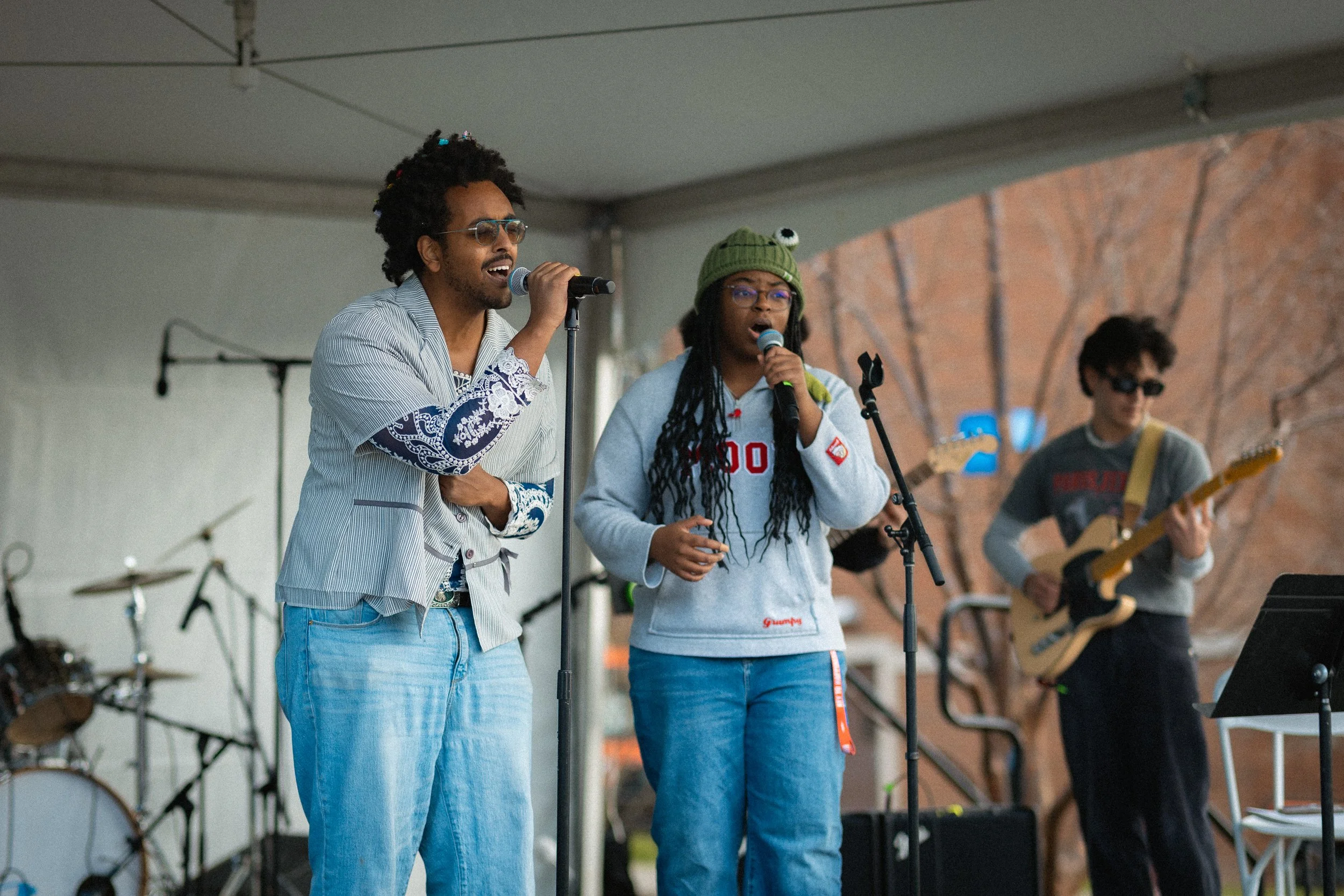 Group of young musicians performing on an outdoor stage, with two singers at microphones and a guitarist in the background.