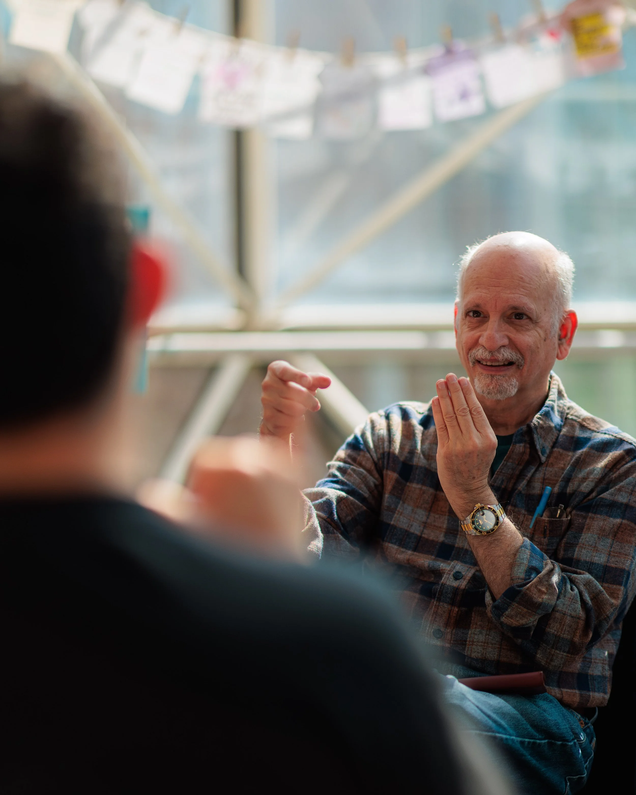 An elderly man with a beard, wearing a plaid shirt and a watch, is smiling and gesturing with his hands. The photo appears to be taken in a casual indoor setting, with a blurred background of hanging papers or photos on a string.
