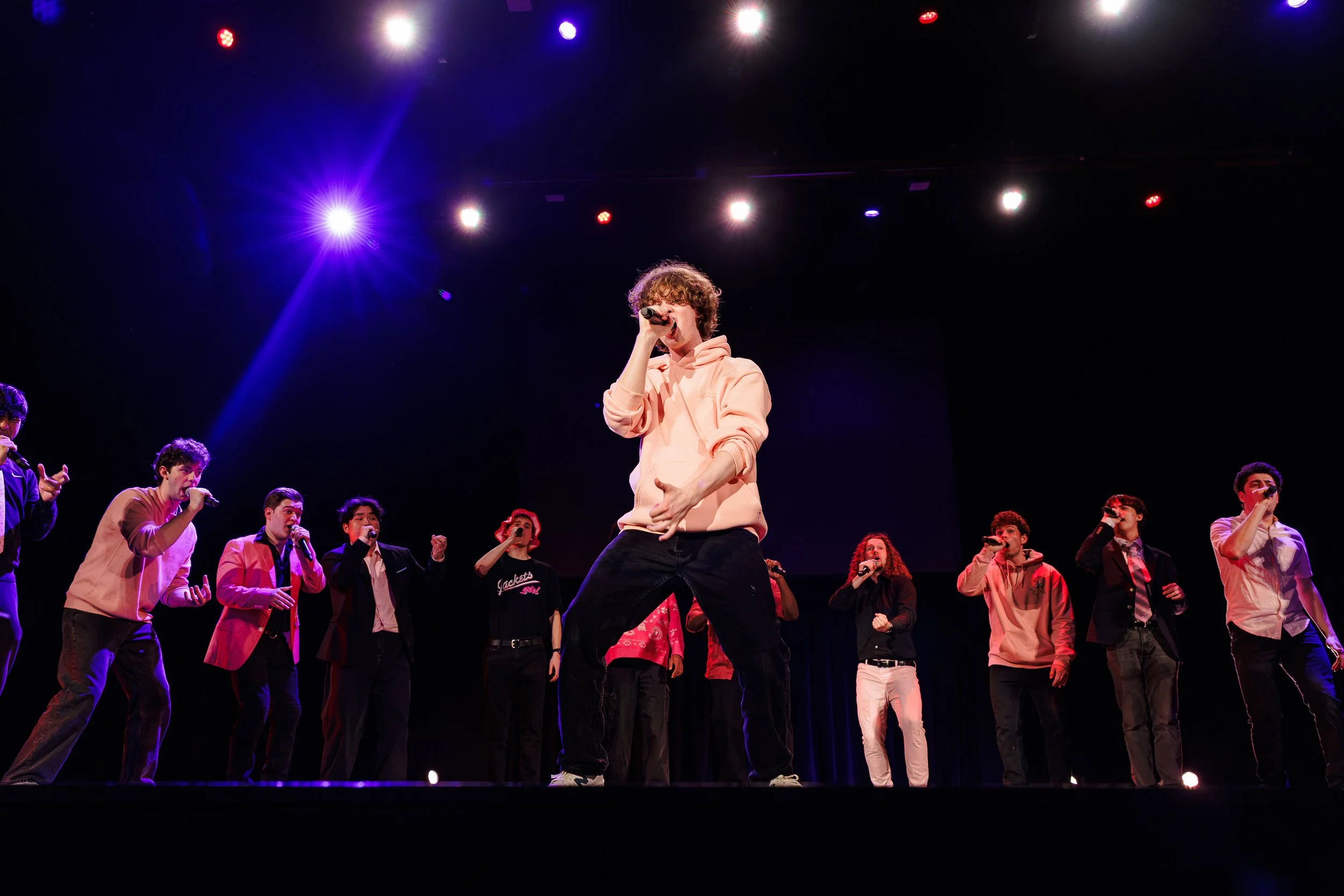 A group of young performers on stage, with one person in the center singing into a microphone. The stage is illuminated with colorful bright lights, and the performers are dressed in casual and semi-formal attire.