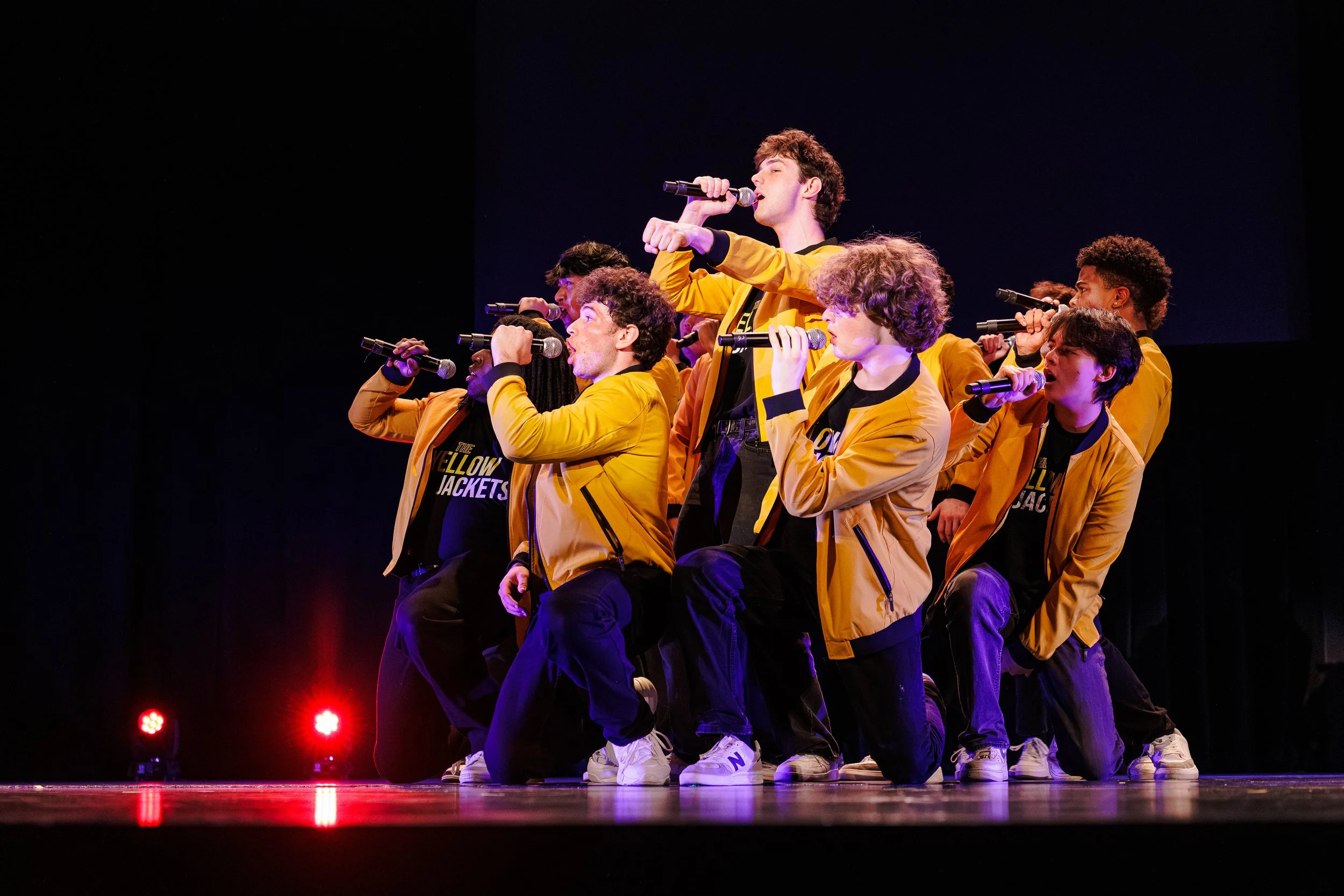 A group of young dancers wearing yellow jackets and black shirts performing on stage with microphones, some kneeling and others standing, with red stage lights in the background.