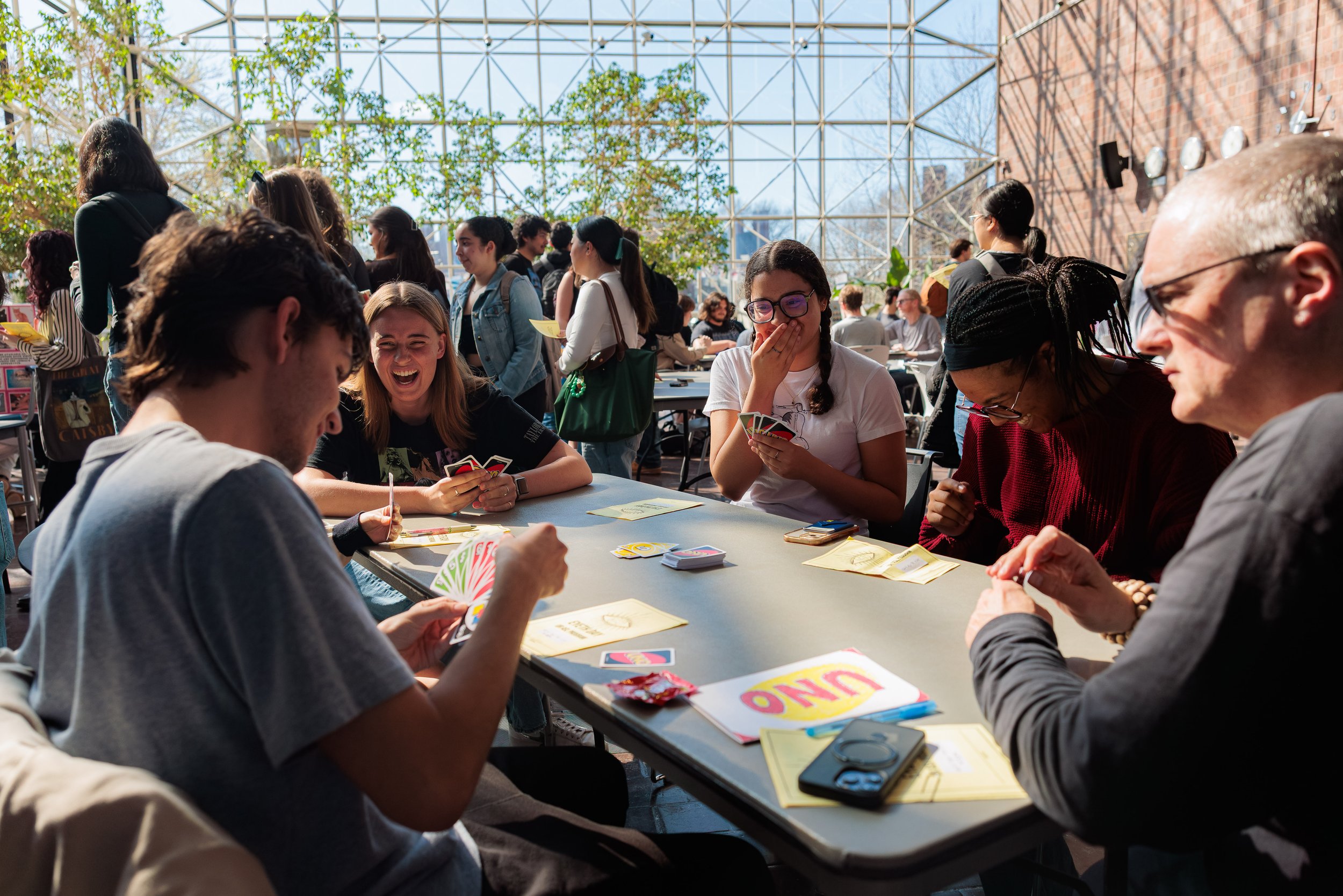 People gathered around playing cards at indoor event with glass ceiling and plants.