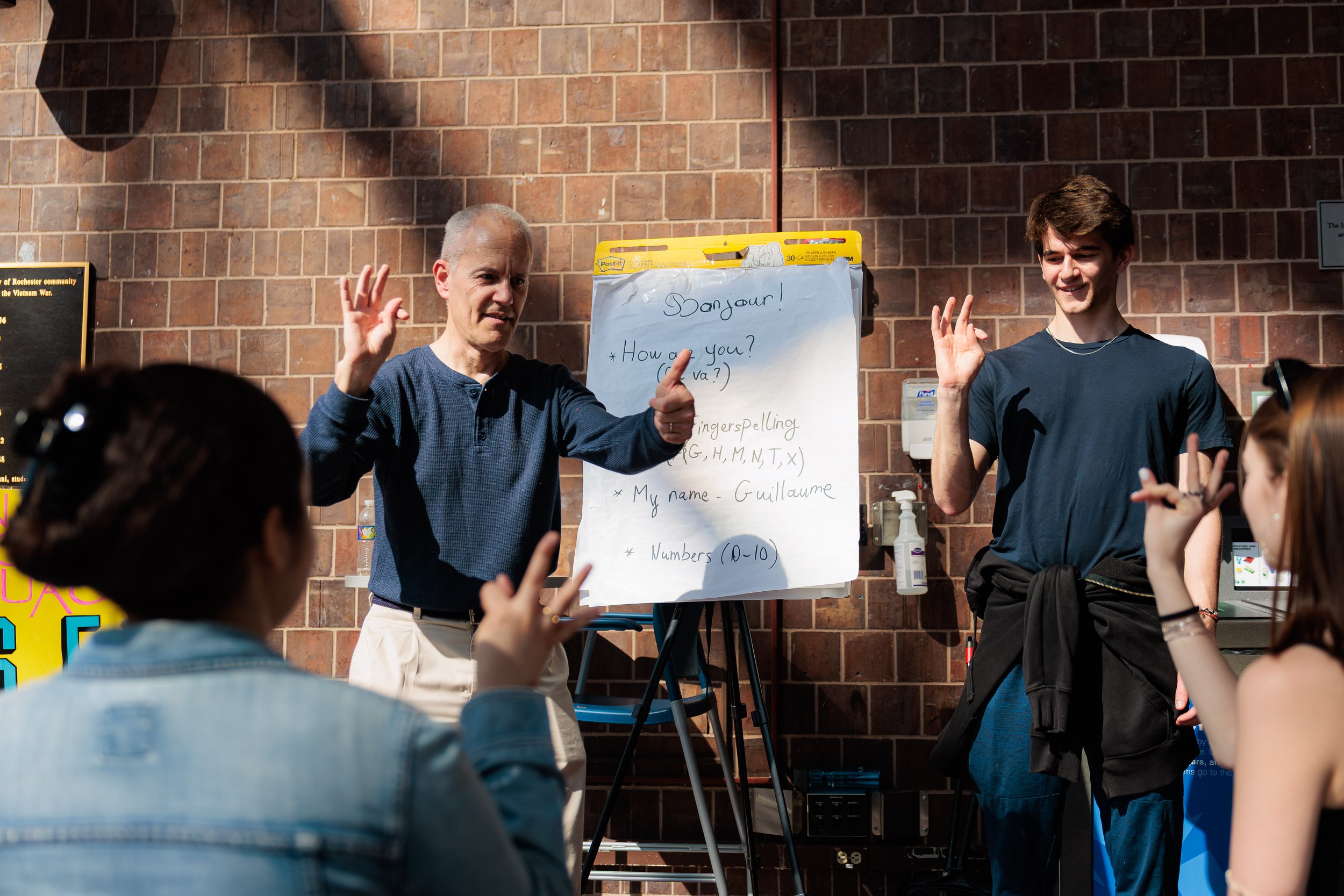A classroom or group setting with a brick wall background, where two individuals are leading an activity. The older man on the left is making an OK hand gesture and the younger man on the right is raising his hand with a friendly smile. There is a wh