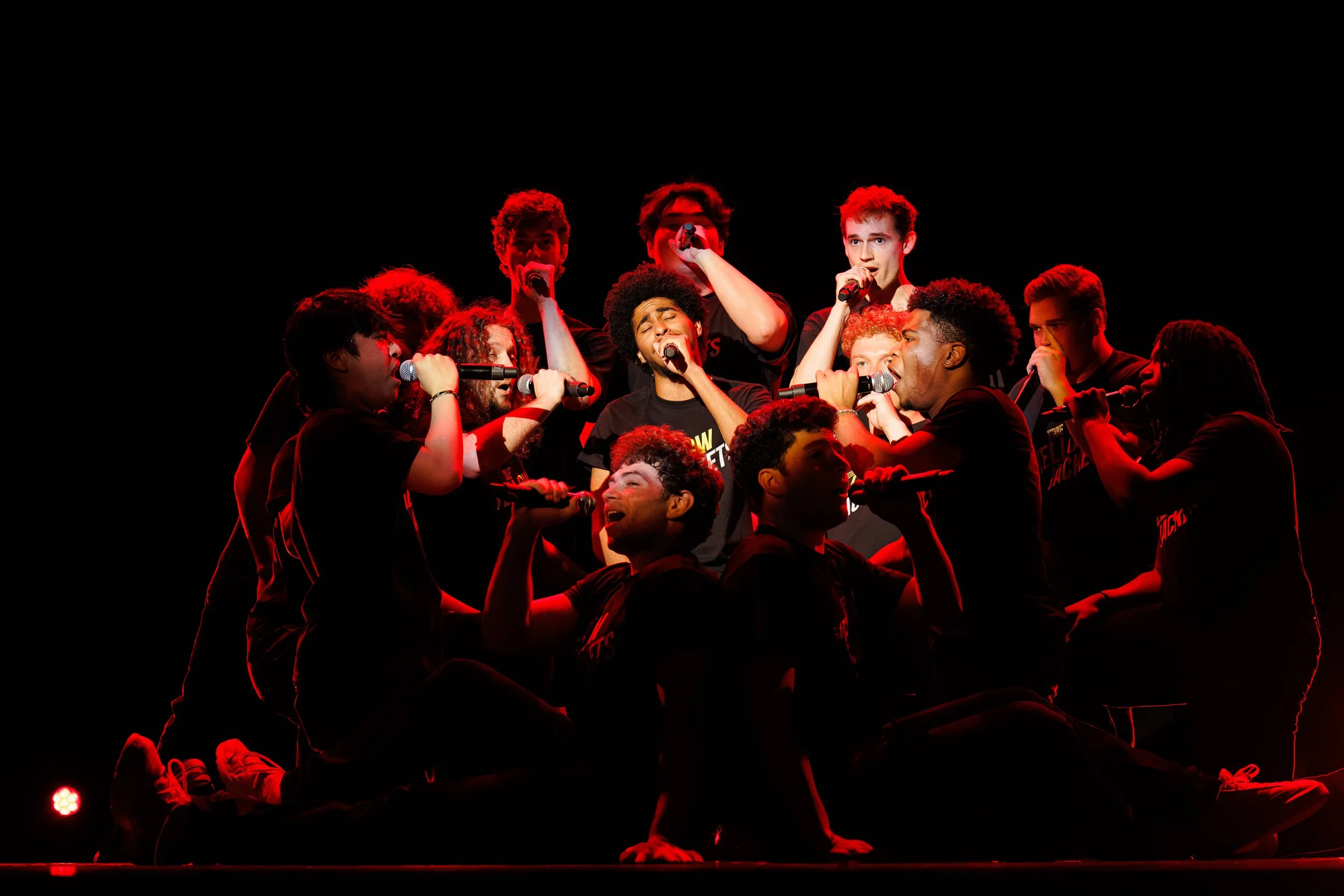 Group of performers singing on stage under red lighting, with some kneeling and others standing, holding microphones.