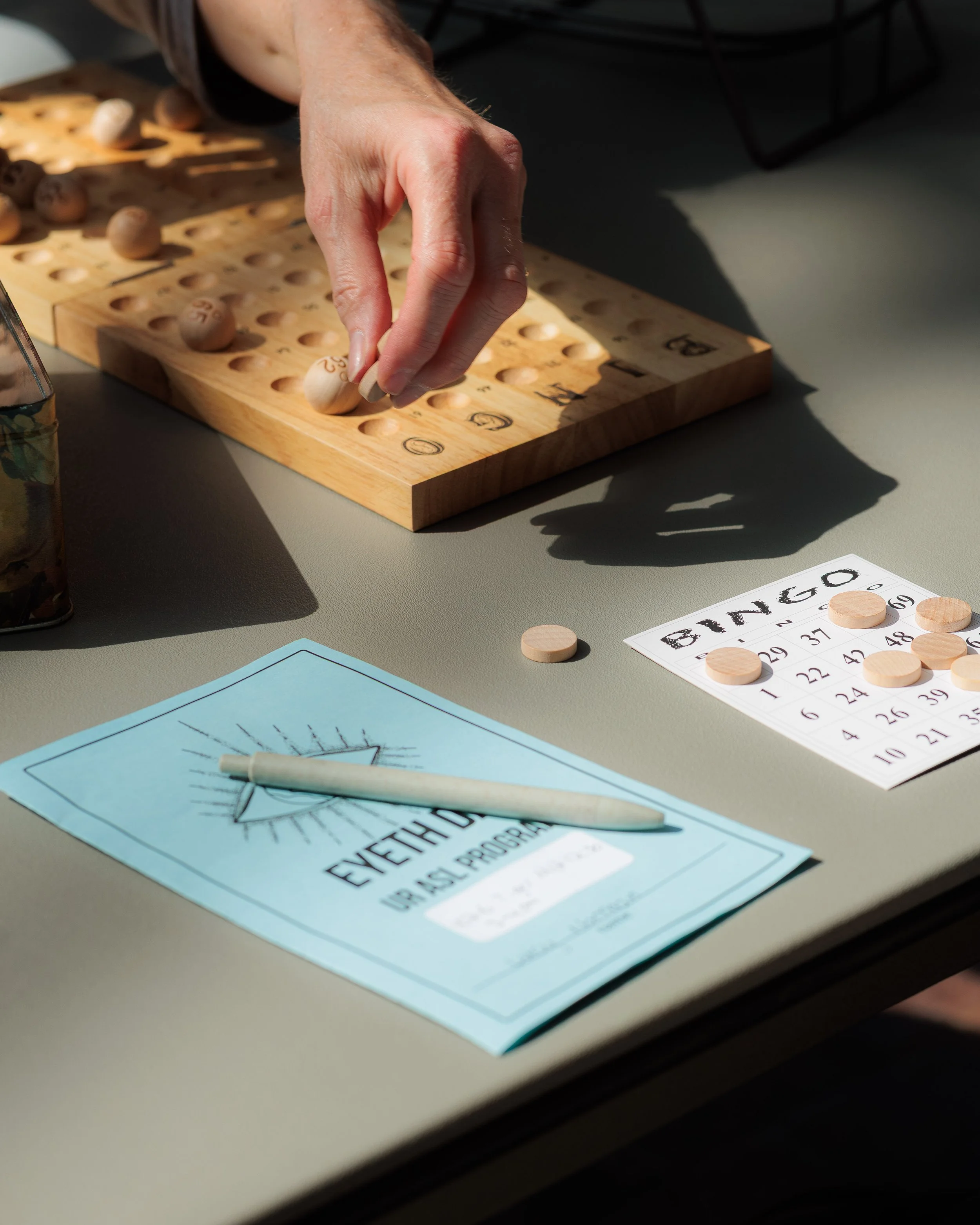 Close-up of a person's hand playing bingo on a wooden bingo board with numbered wooden balls, a bingo card on the table, and a blue flyer with a sun illustration.