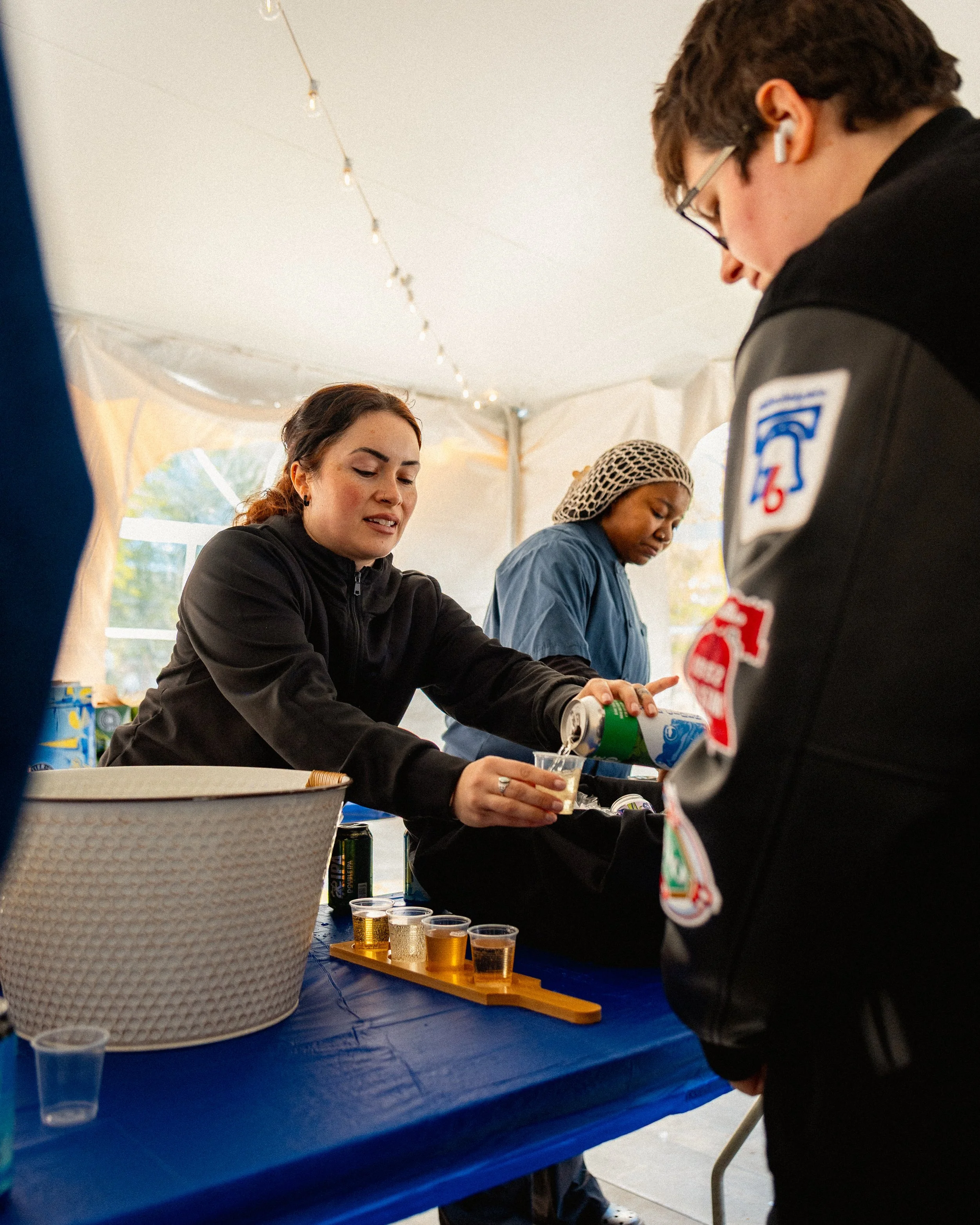 People pouring and drinking beer at an outdoor event under a tent, with string lights hanging above.