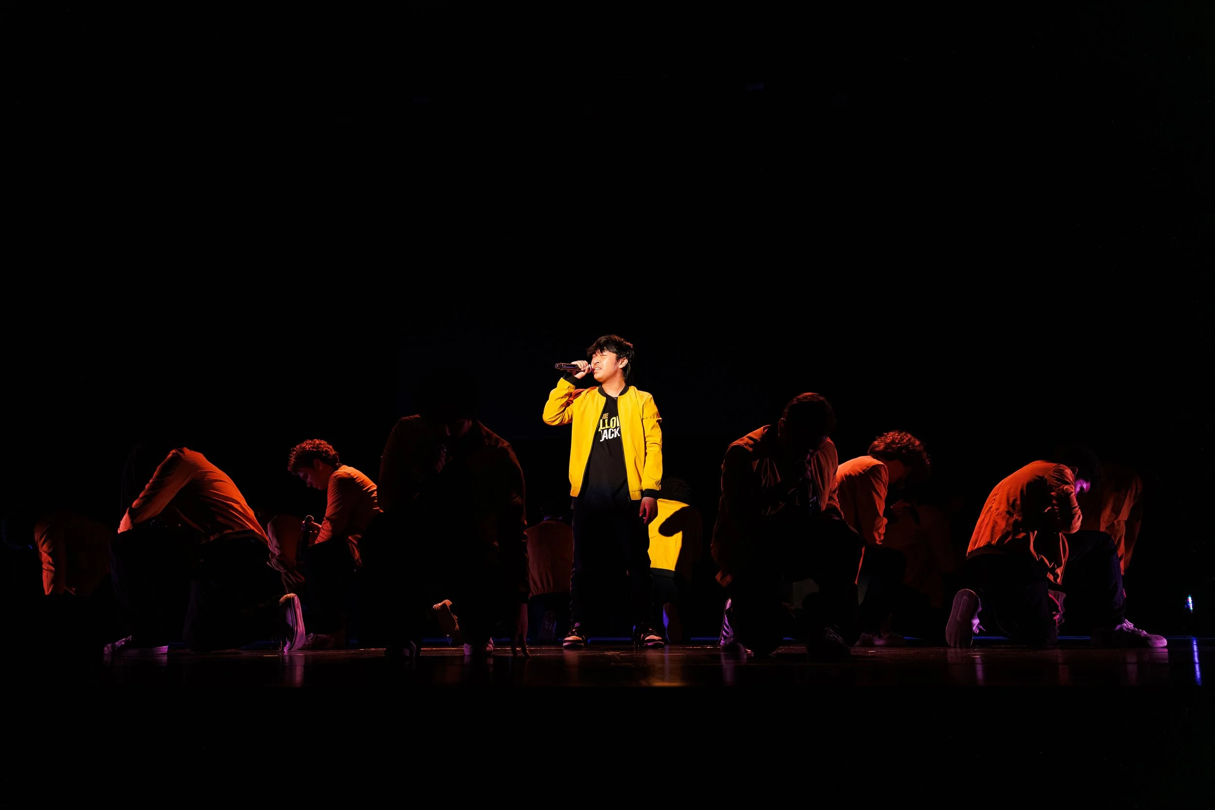 A person in a yellow jacket standing on stage, singing into a microphone, with a group of people kneeling or bowing in front of them under a dark background.