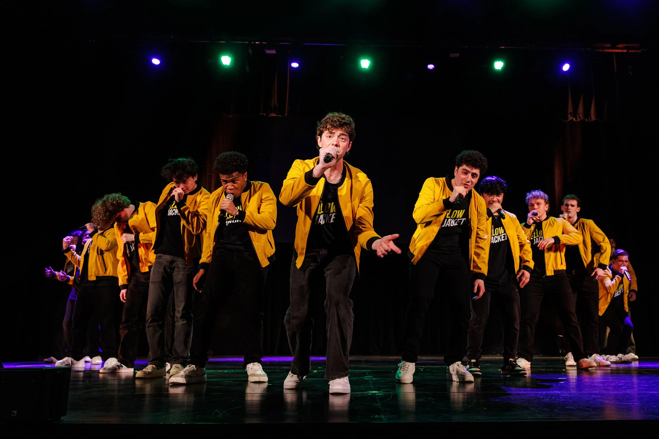 A group of young performers on stage, wearing yellow jackets, singing into microphones during a concert or talent show, with stage lighting overhead.