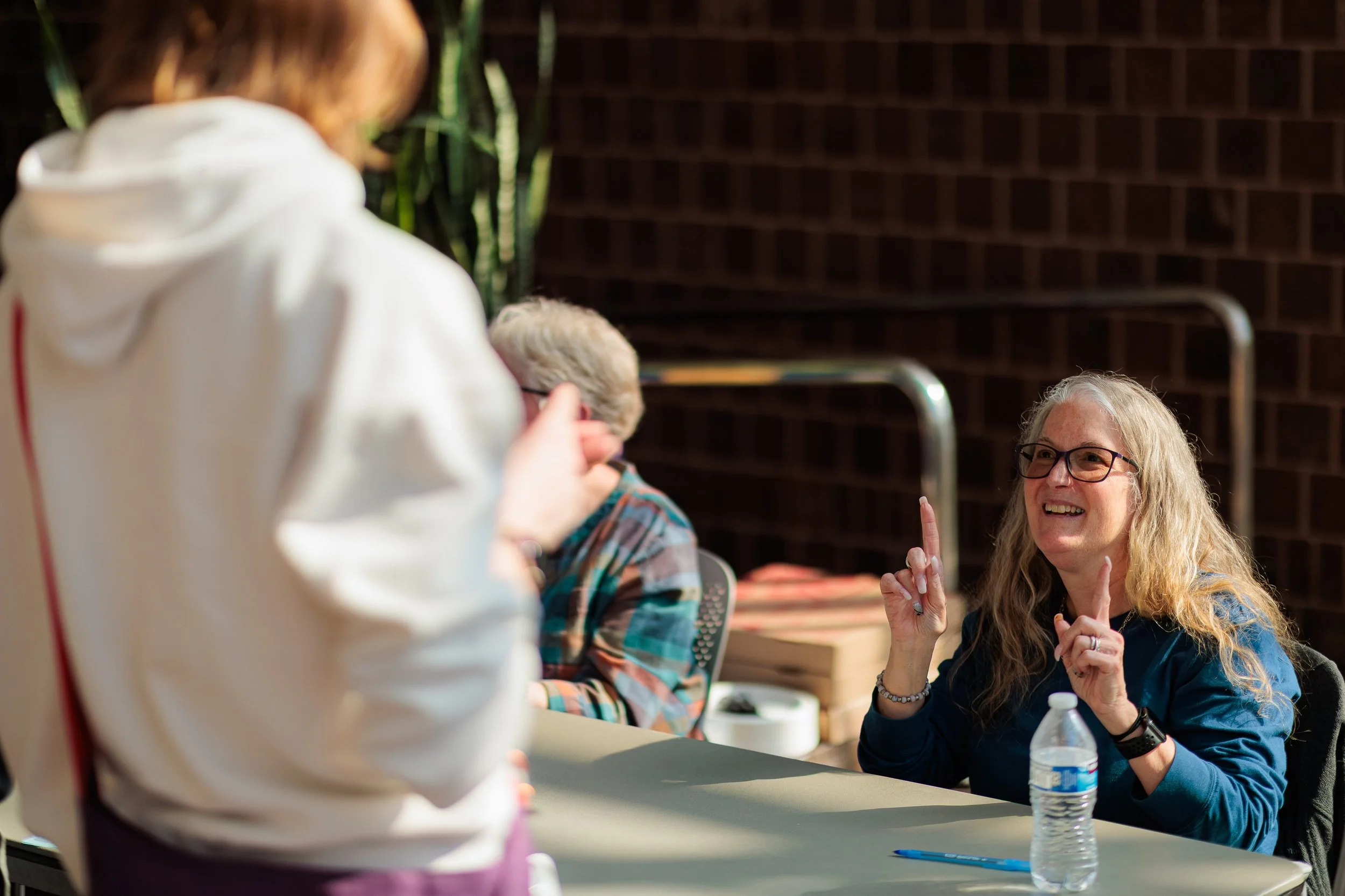A woman with glasses and long blonde hair sitting at a table, smiling and raising her hands with two fingers up, while talking to a person in front of her. The person in front has light-colored hair and is wearing a white hoodie. There is a water bot