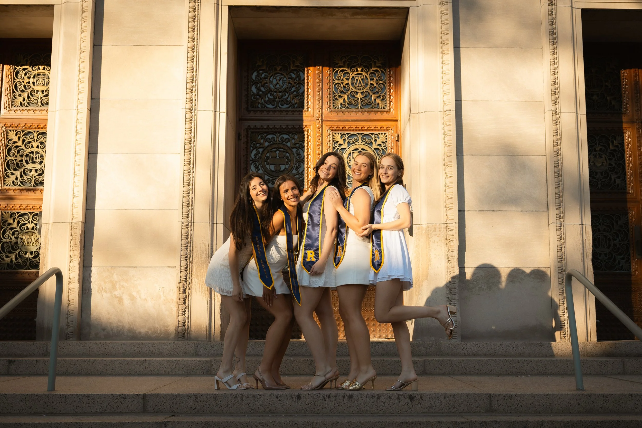Group of five young women in elegant dresses and high heels, wearing graduation sashes, standing on steps in front of a decorative building with ornate wooden doors and columns, smiling for a photo.