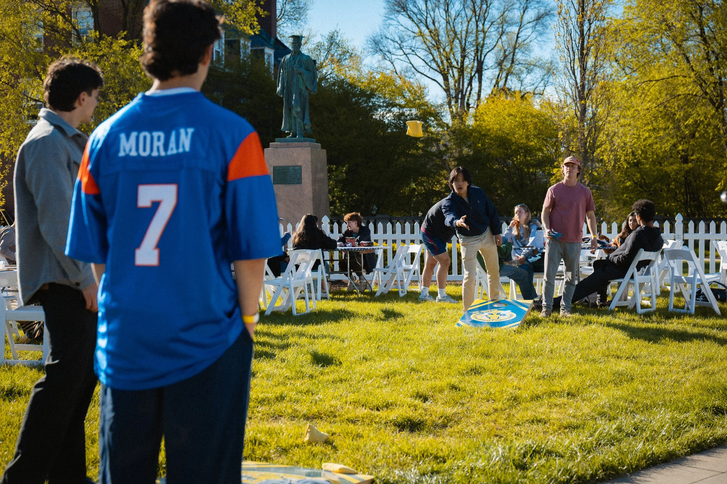 Two boys, one wearing a blue and orange jersey with the name 'MORAN' and number 7 on the back, stand outside on a grassy area watching a game. In the background, people are sitting on white chairs and some are playing cornhole, with a statue of a his