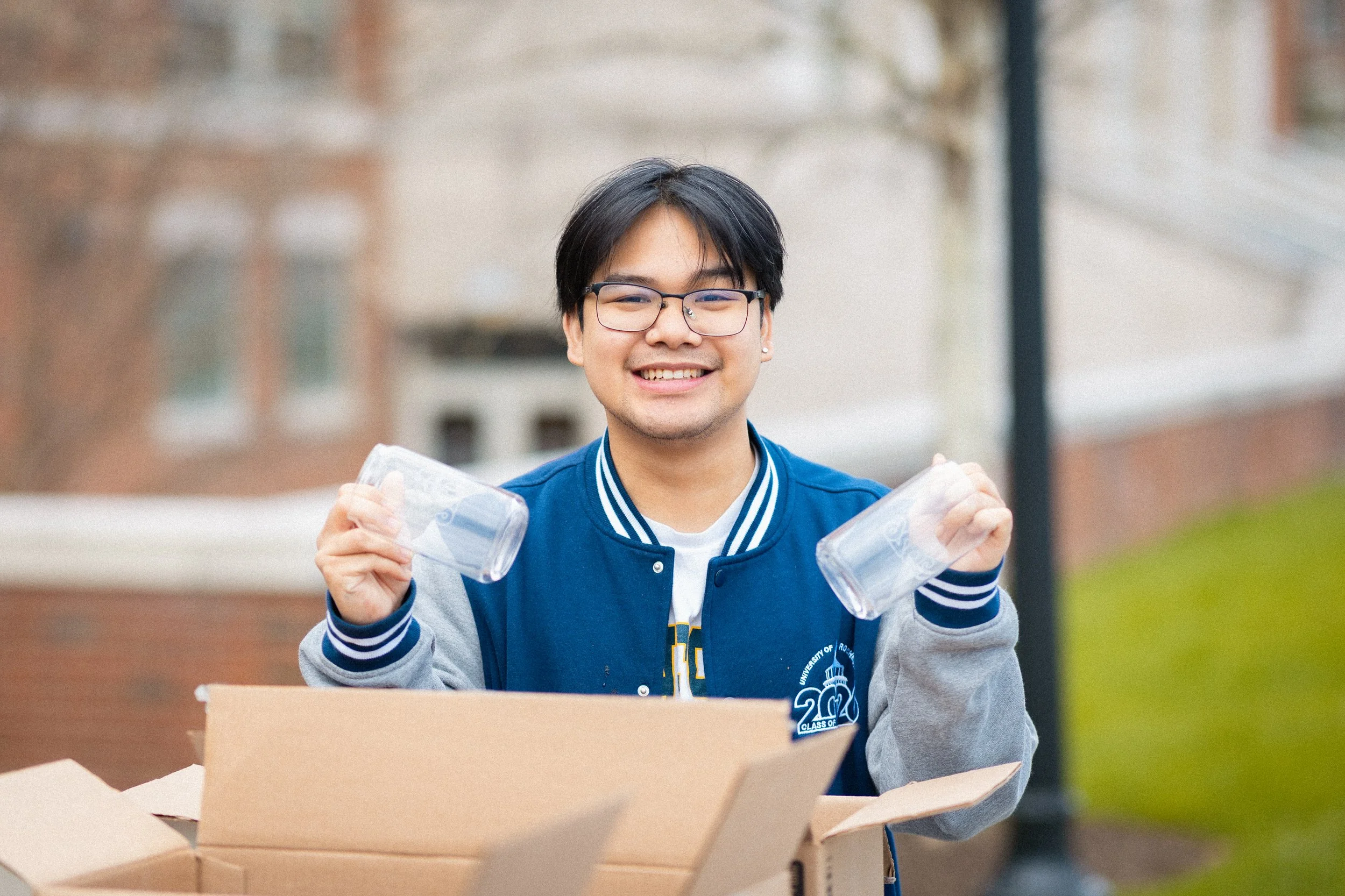 A young man with glasses and dark hair smiling, holding two clear plastic cups, standing behind an open cardboard box outdoors with blurred buildings and trees in the background.