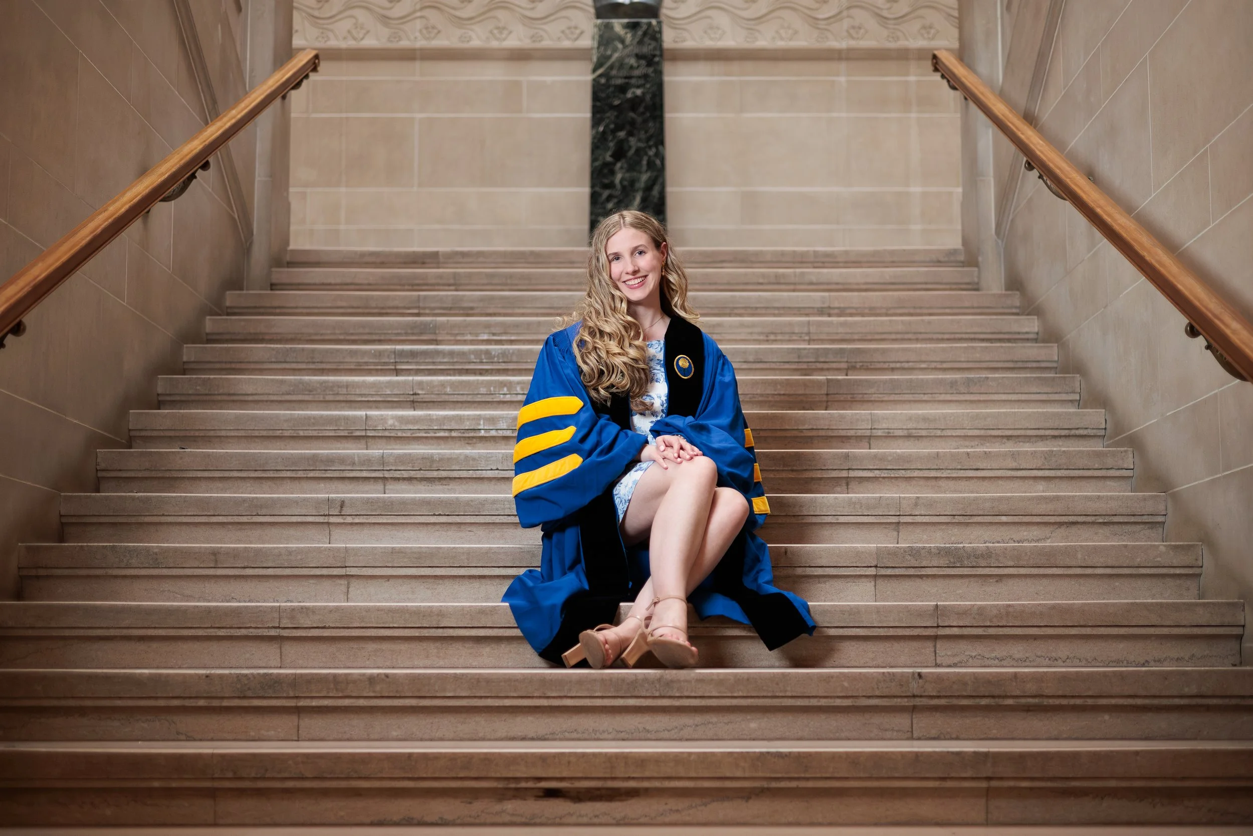 Young woman in a blue graduation gown sitting on stairs with a smile.