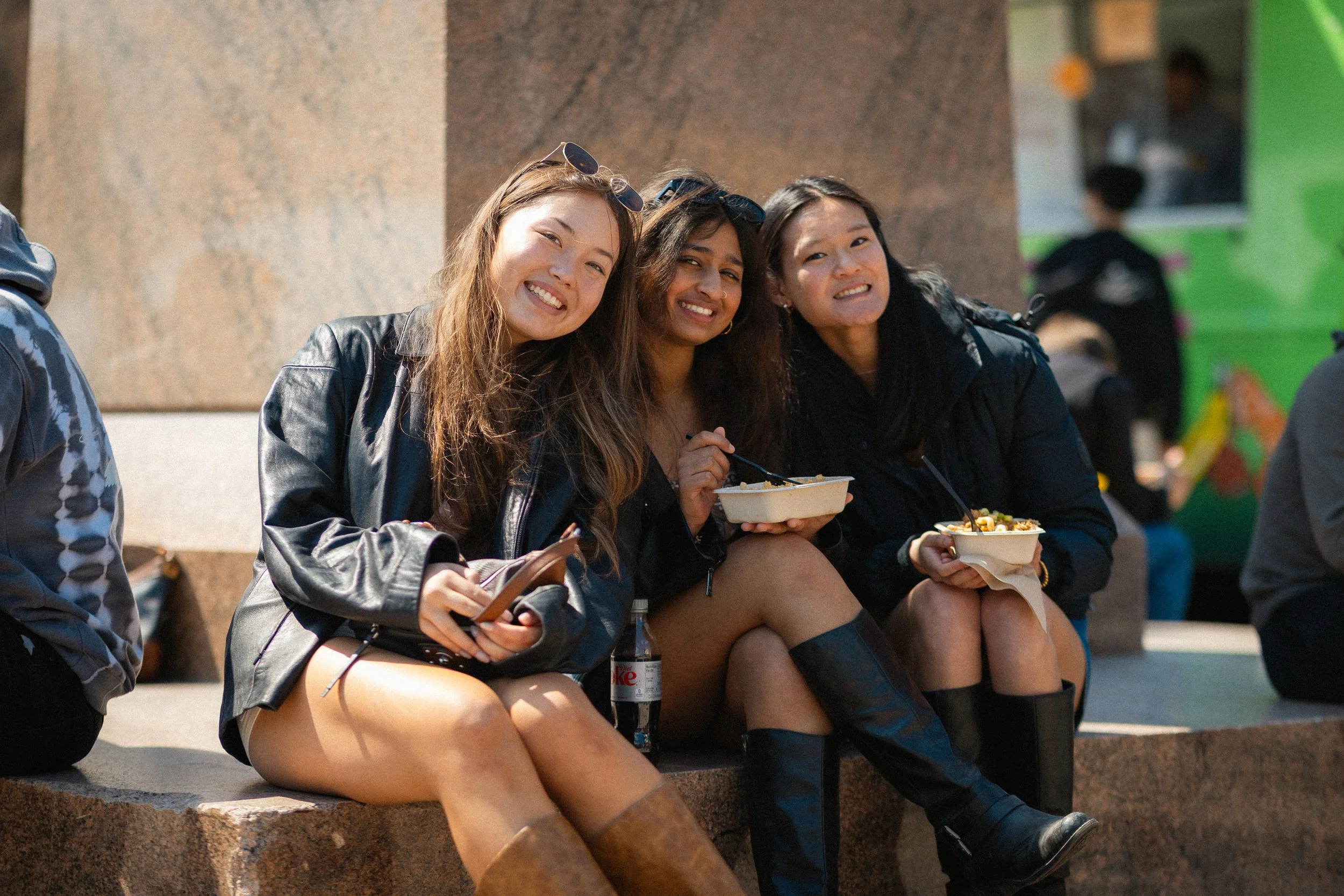 Three young women sitting on steps outdoors, smiling and eating food from takeout containers, dressed in casual clothing and black boots.