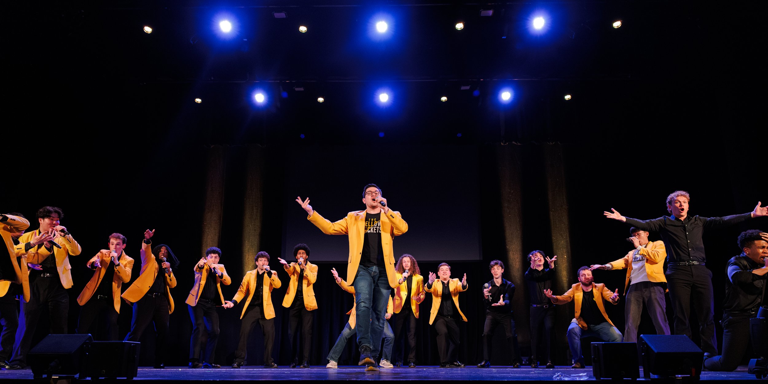 A group of performers, mostly wearing yellow jackets, on a stage with a dark background and bright stage lights, singing or speaking into microphones.