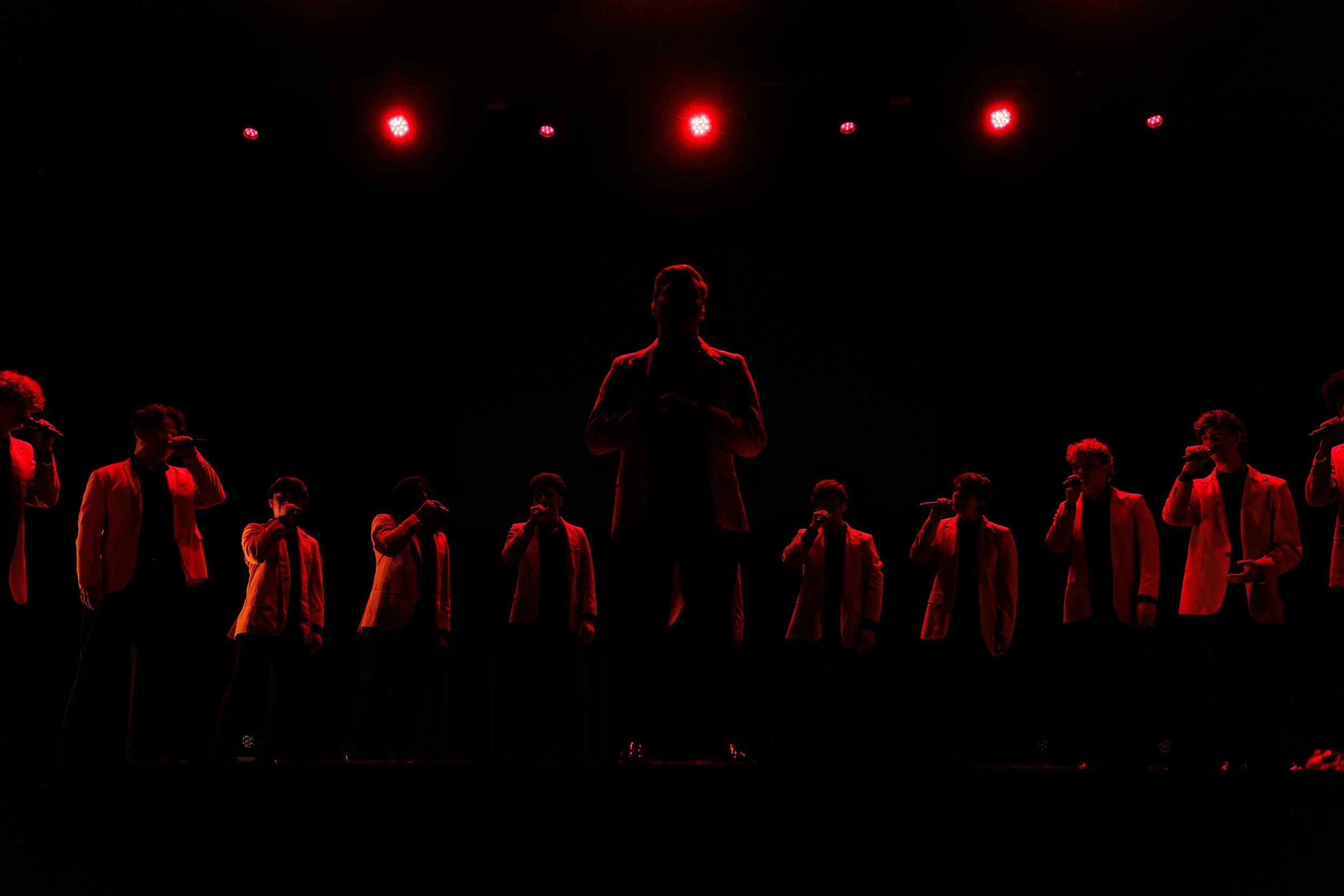 A performer standing on stage illuminated by red lights, surrounded by other performers also holding microphones, all wearing matching outfits.