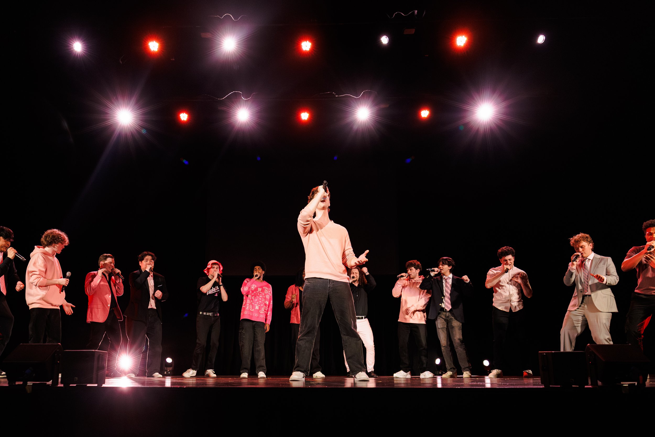 Group of young people on stage performing with microphones, some wearing pink and casual attire, and one person in the center holding a microphone with a spotlight above.