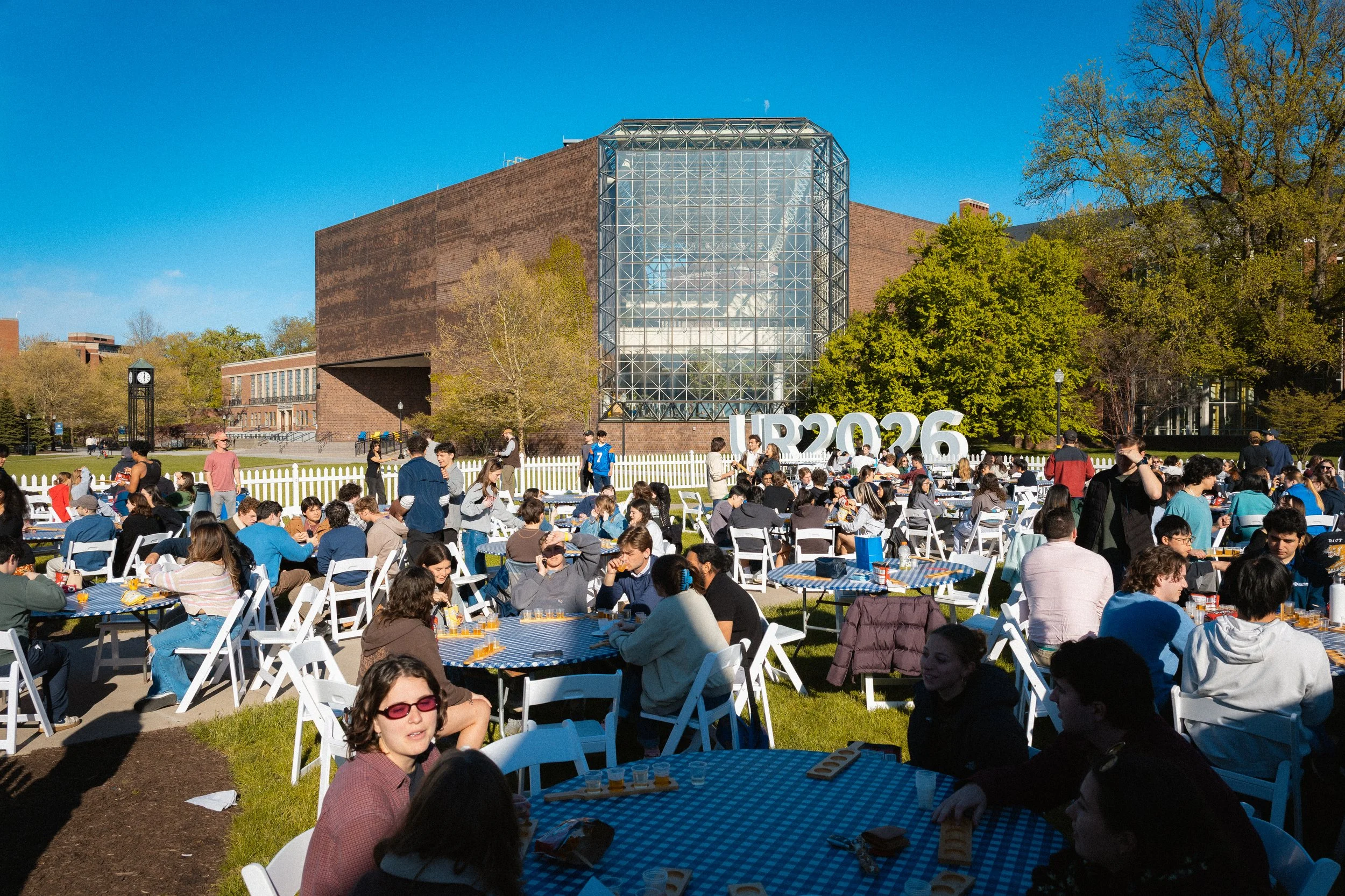 Outdoor graduation celebration with numerous people seated at tables with blue checkered tablecloths, a large brick building with glass and steel structure, green trees, and a bright blue sky.