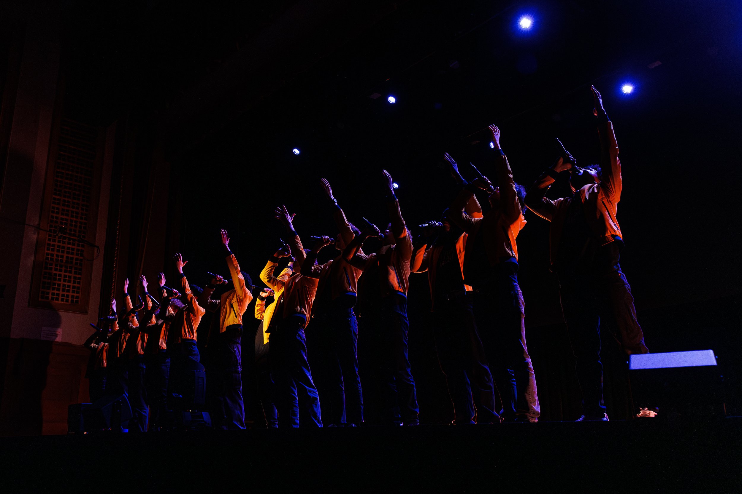 A group of people standing on stage with their arms raised, illuminated by stage lights in a dark theater.
