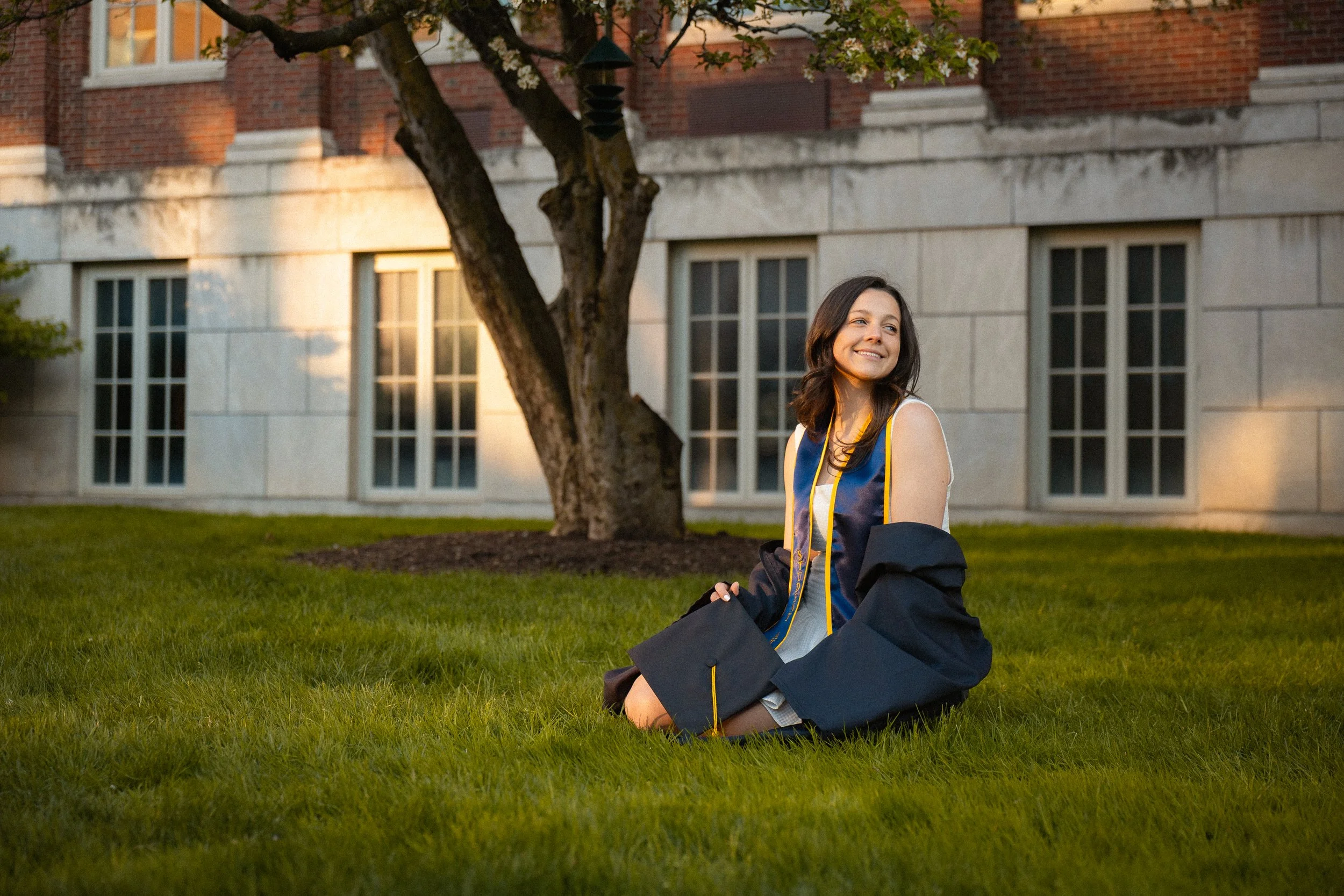 A young woman in graduation attire sitting on the grass in front of a brick building and tree, smiling after graduation.