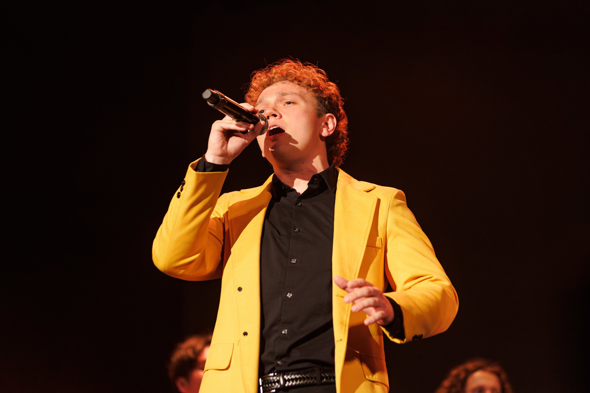 A man with curly hair singing into a microphone on stage, wearing a yellow blazer and black shirt.
